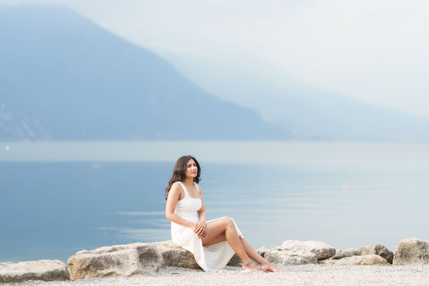 Someone in a white dress sits on coastal rocks overlooking a misty lake and mountains in the background.