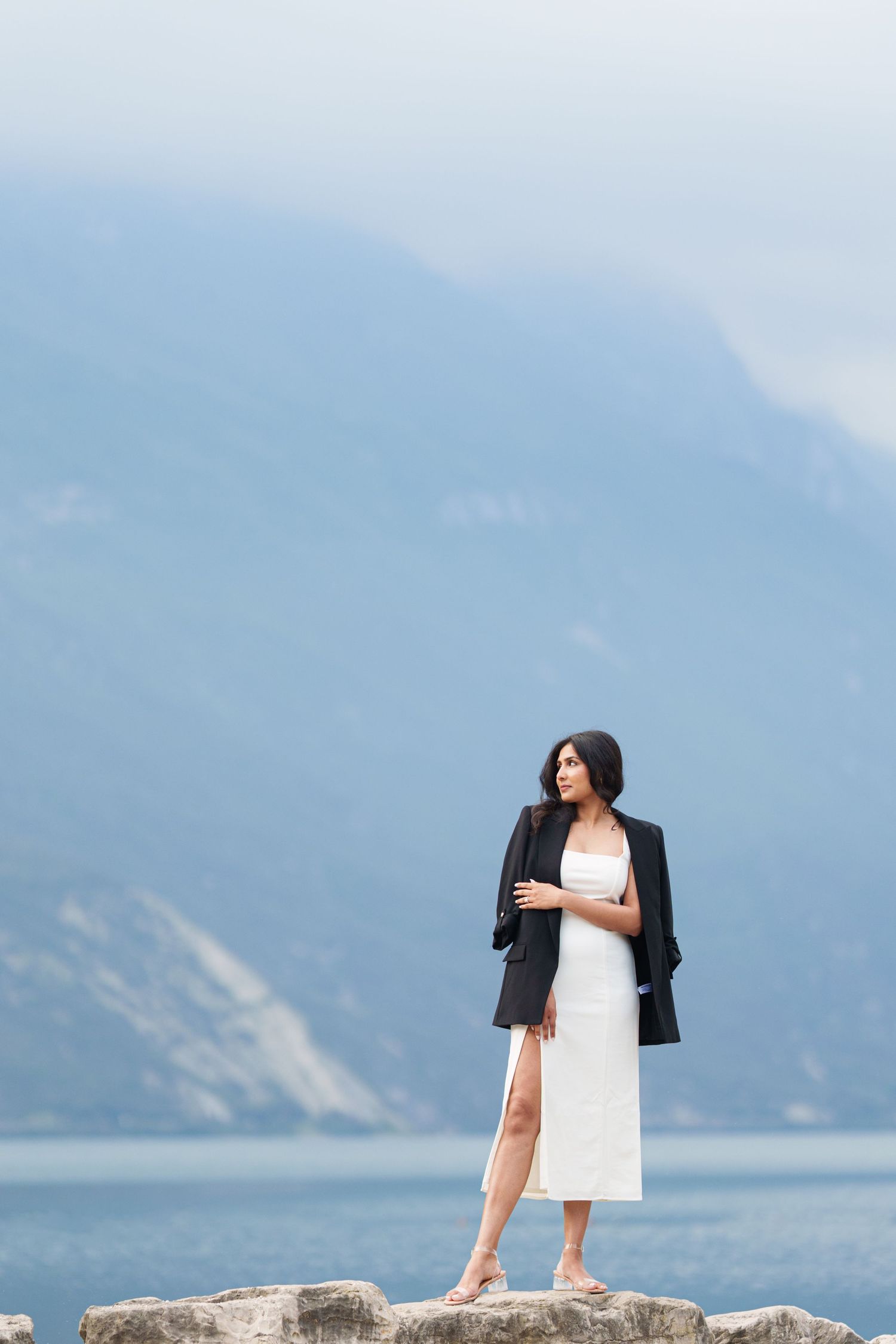 A person in a white dress and black blazer stands on a rocky overlook with mountains and a lake in the background.