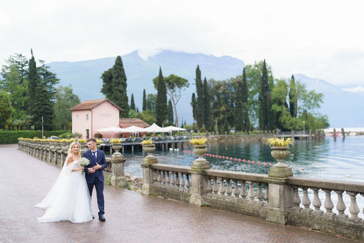 A couple walks along a scenic lakeside promenade with mountains and cypress trees in the background.