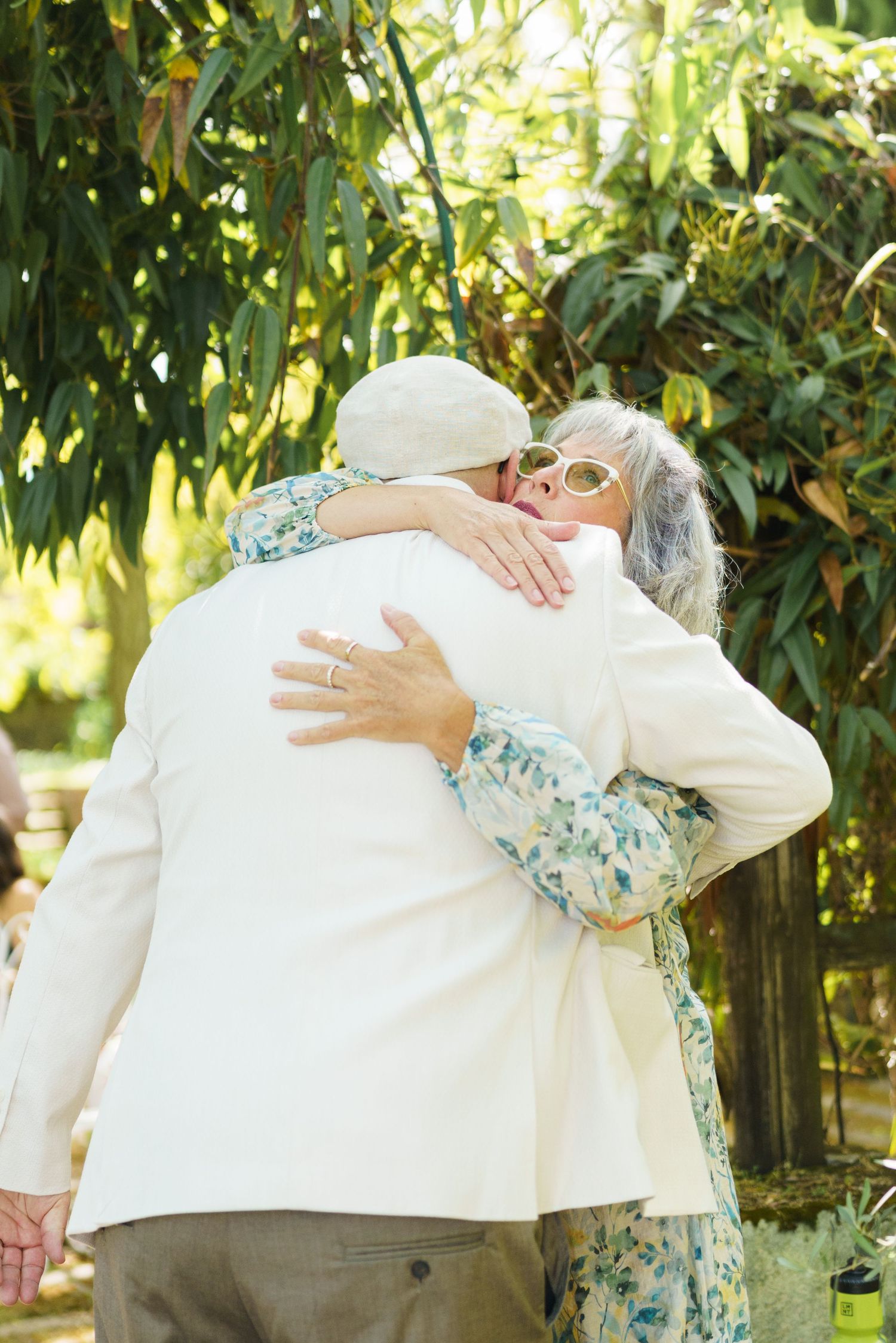Elderly couple sharing a warm hug outdoors, surrounded by lush green foliage on a sunny day.