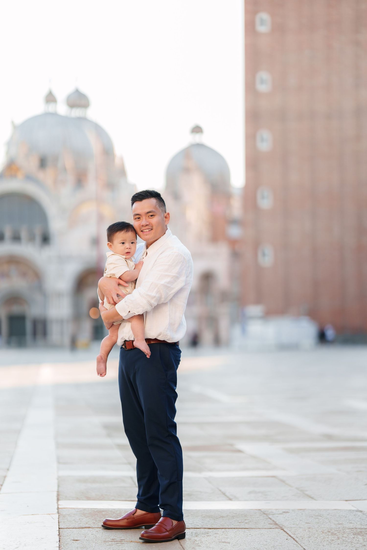 A smiling man holds a baby in St. Mark's Square, Venice, with the iconic basilica in the background.