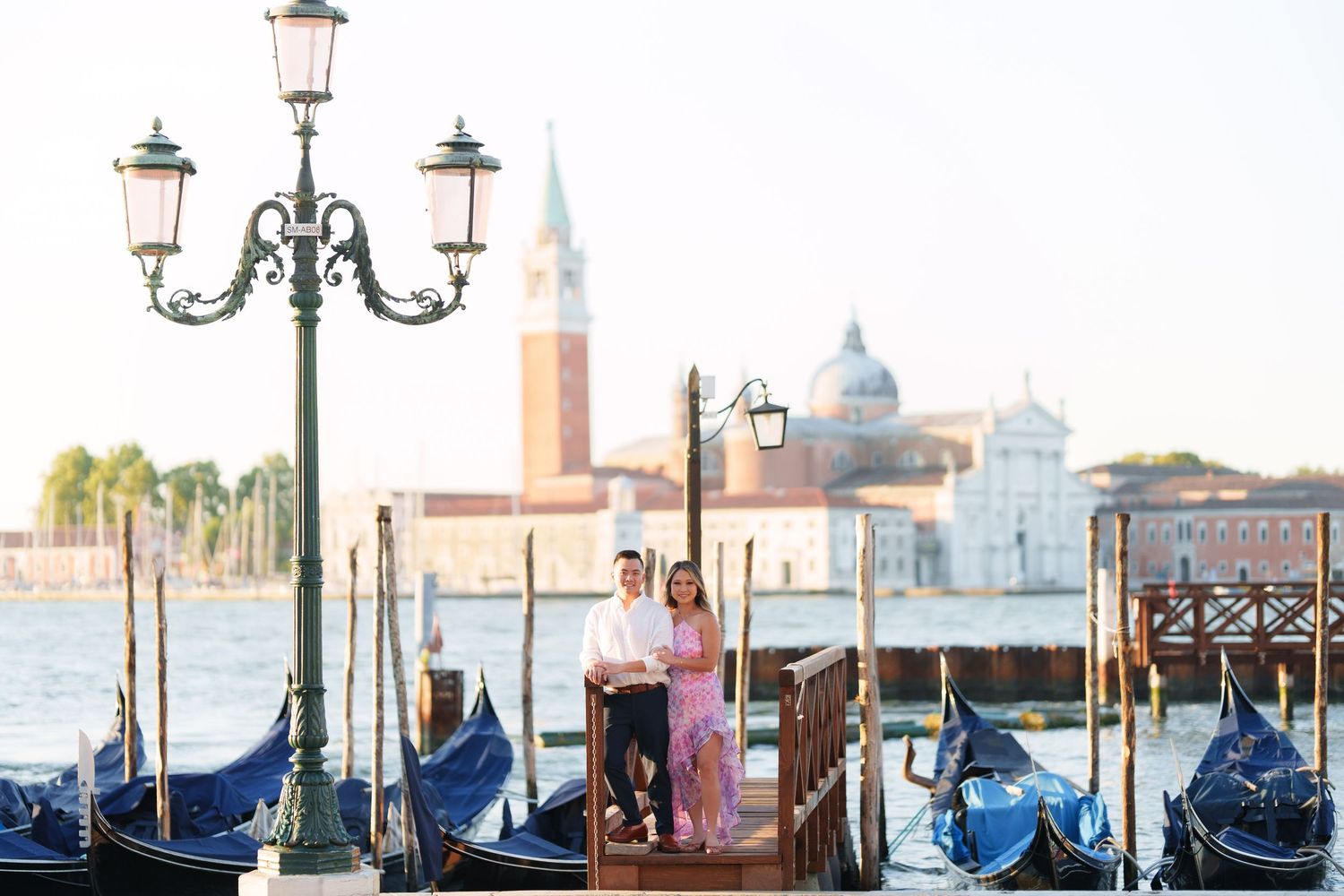 Couple standing on Venice gondola dock with San Giorgio Maggiore church and grand canal in soft morning light.