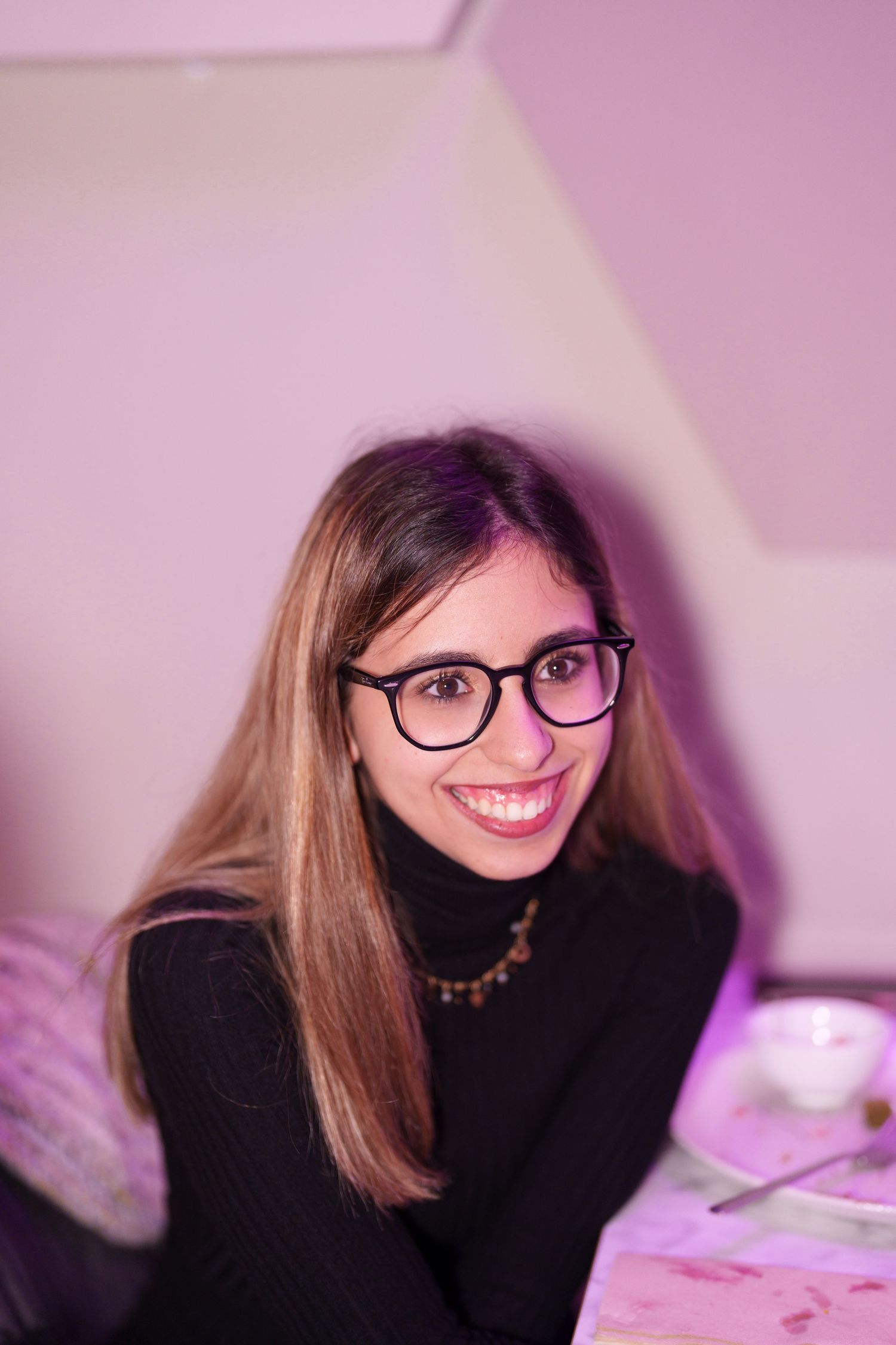 Young woman with long hair and black glasses smiling at a table in a pink-lit room wearing a black turtleneck.