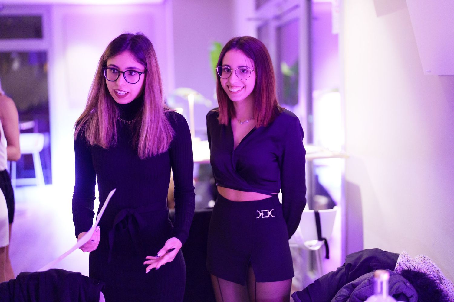 Two young women in black outfits posing together at a purple-lit event venue.
