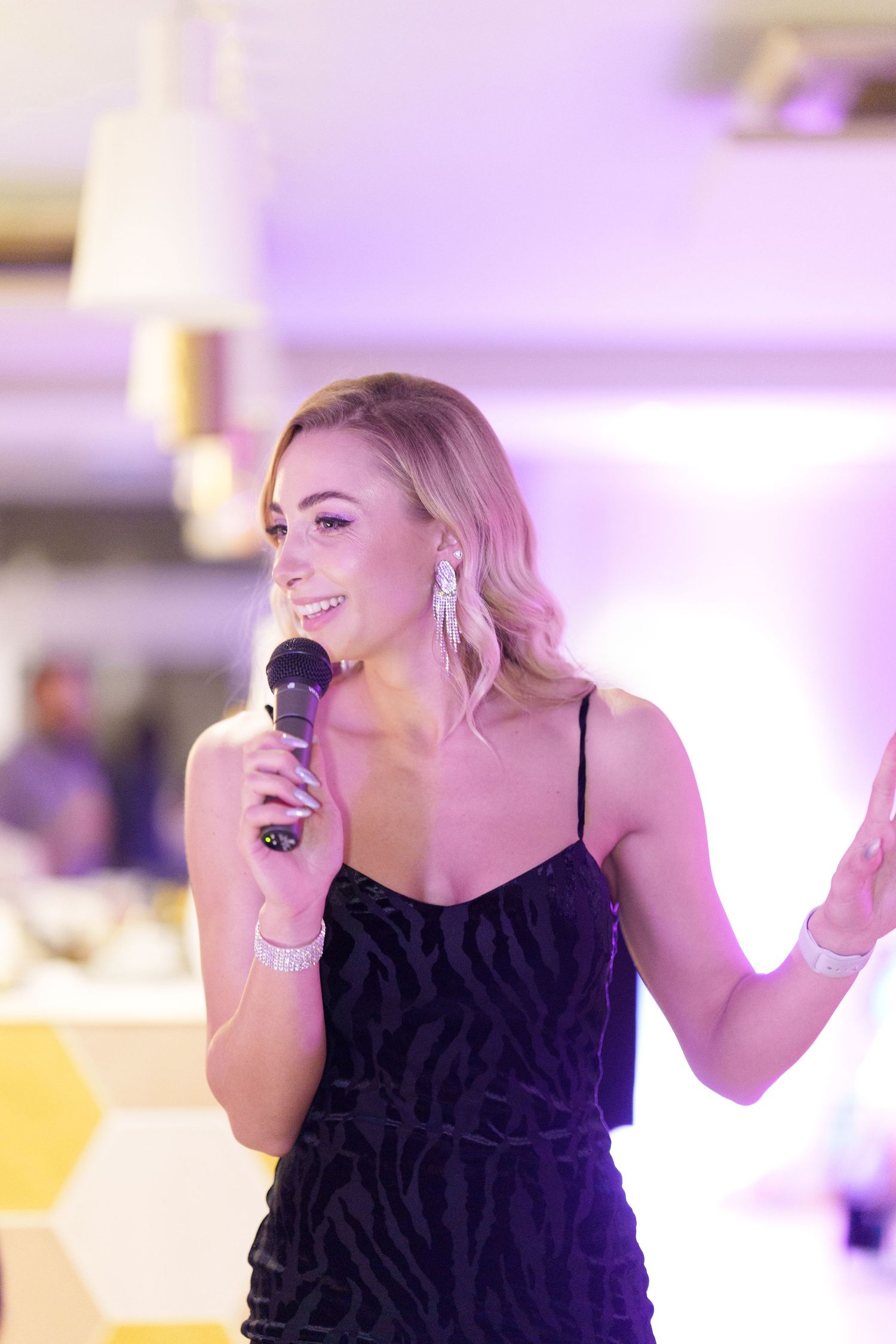 Blonde woman in black dress speaking into microphone at elegant event with purple lighting.