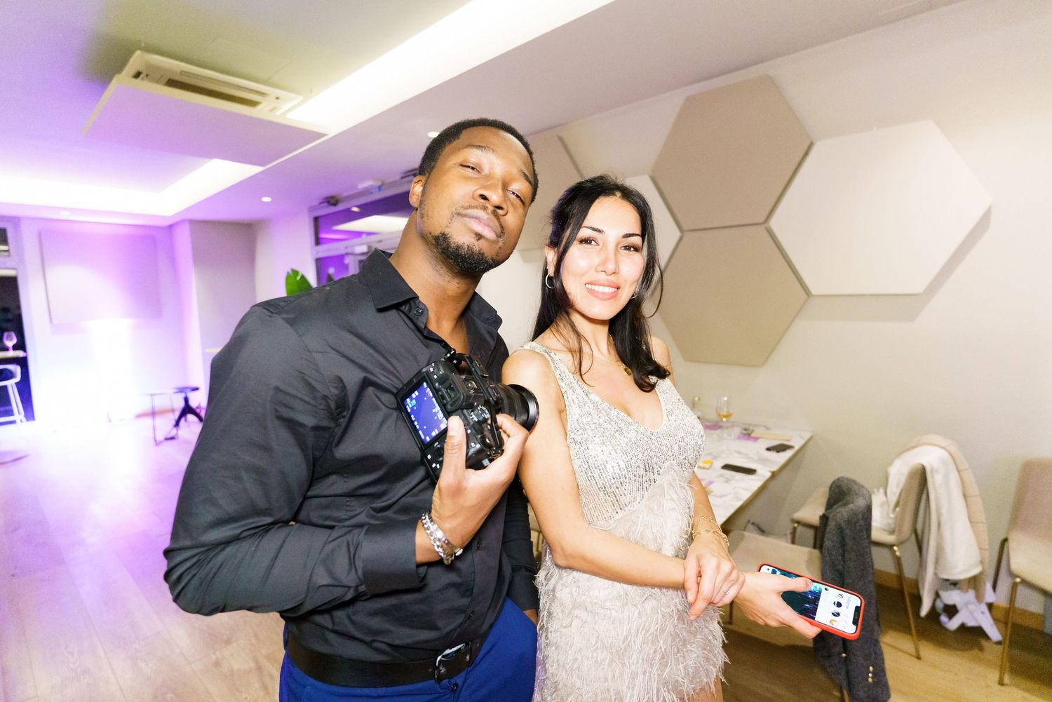 A man holding a camera and a woman in a sequined dress pose together at an indoor event with purple lighting.
