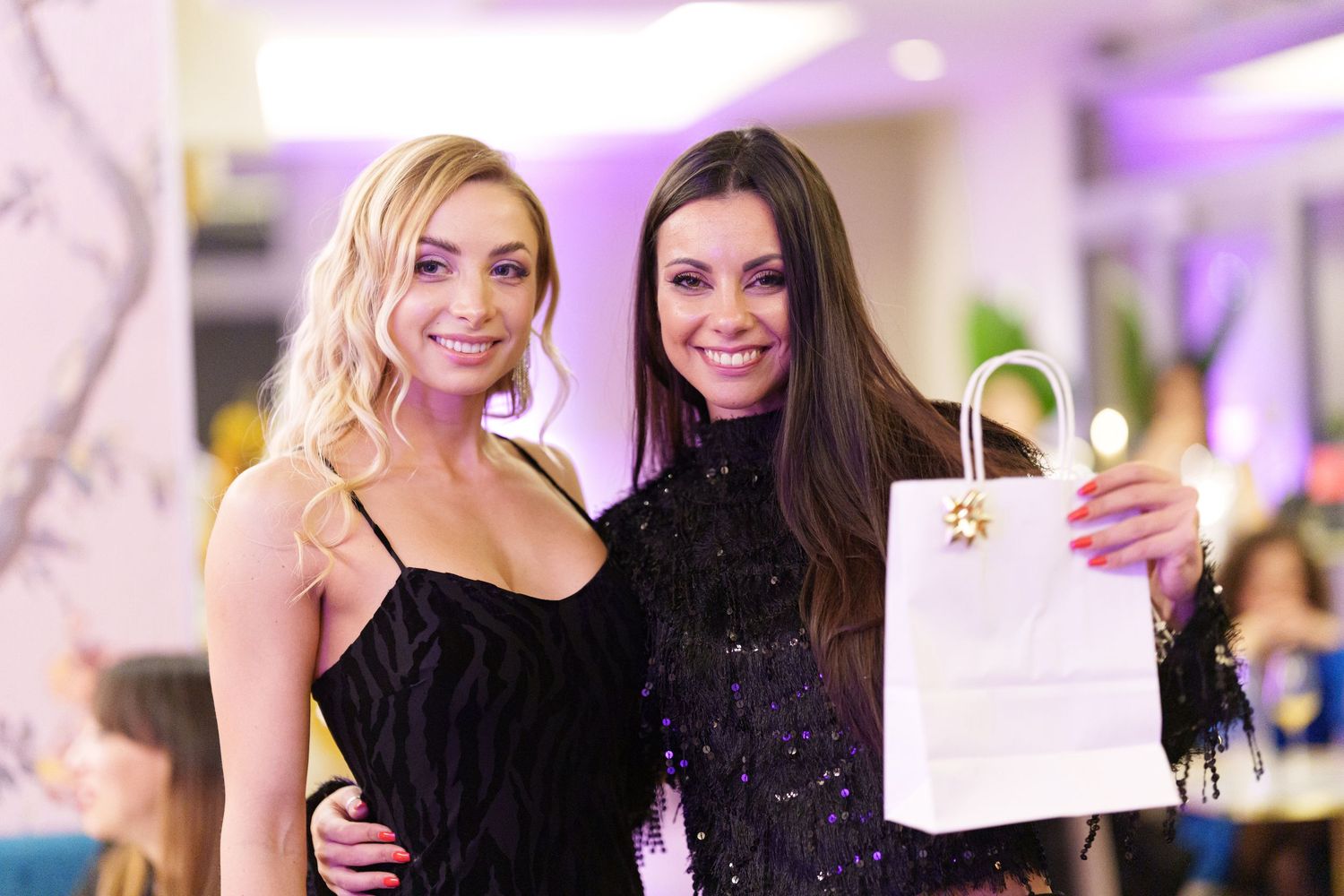 Two elegant women in black dresses posing together at an event, one holding a white shopping bag.