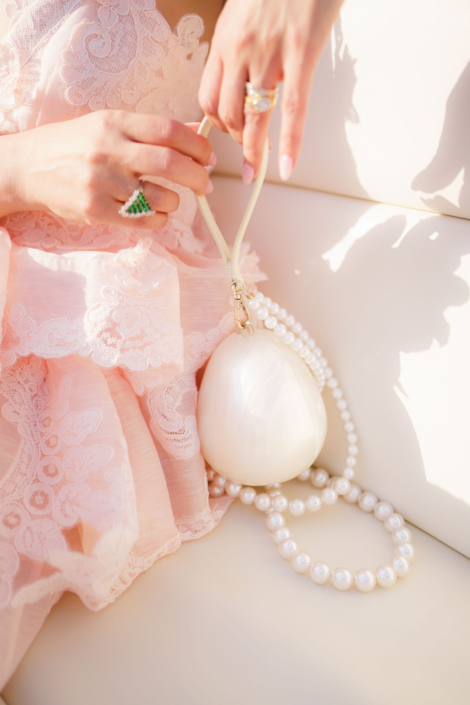 Woman in pink lace dress holding pearl egg-shaped bag with pearl necklace, wearing green gemstone ring.