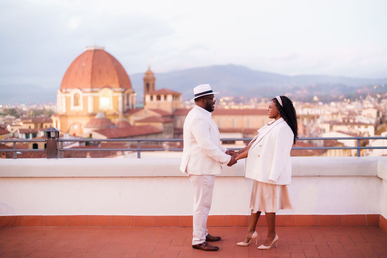 Couple in white outfits holding hands on rooftop with Florence Cathedral dome and cityscape at dusk.