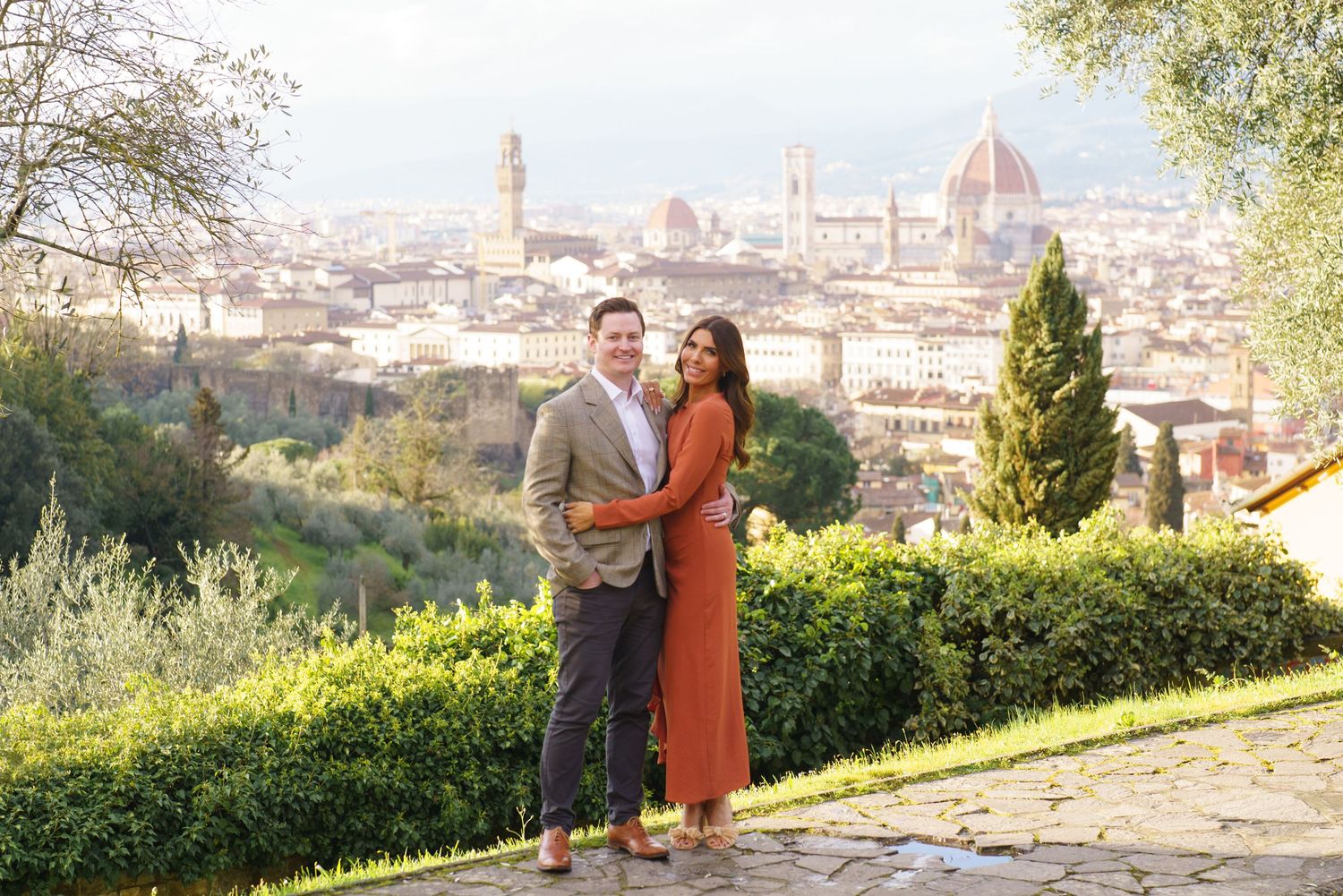 Couple embracing with Florence skyline and cathedral dome backdrop in a lush garden setting.