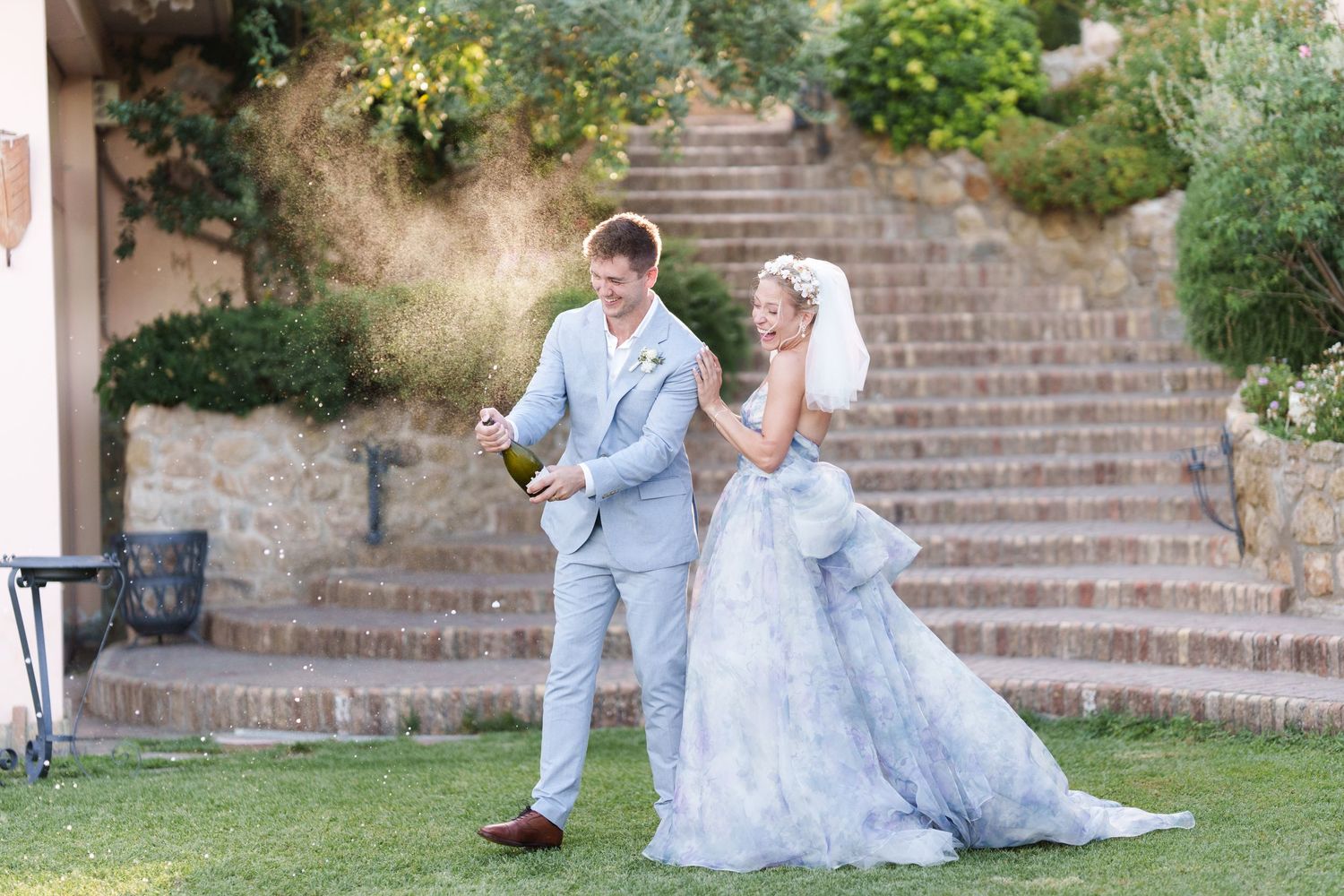 Bride in blue gown and groom in gray suit opening champagne bottle outdoors on green lawn at wedding.
