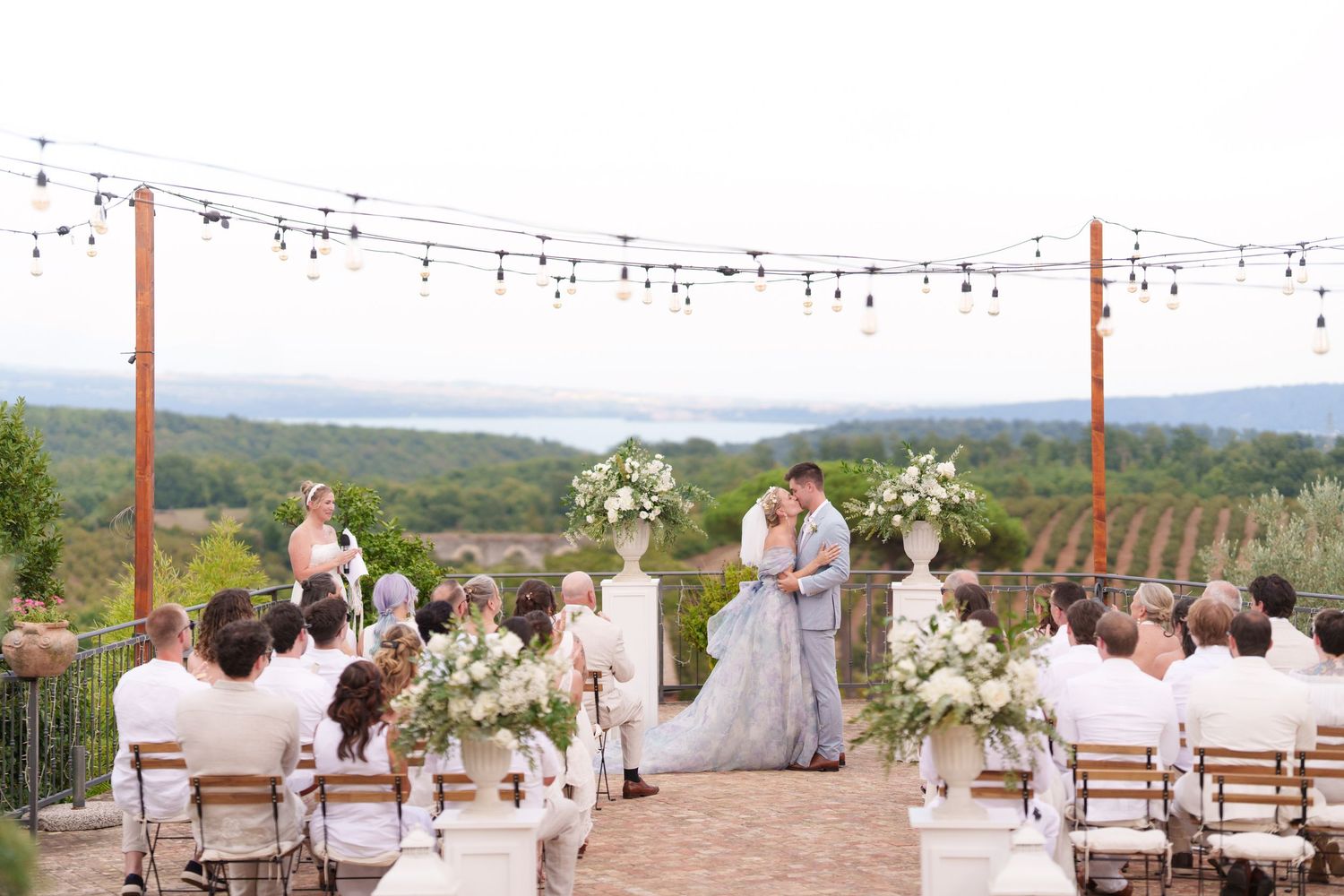 Outdoor vineyard wedding ceremony with couple at floral altar, string lights, white chairs, and scenic misty mountain views.