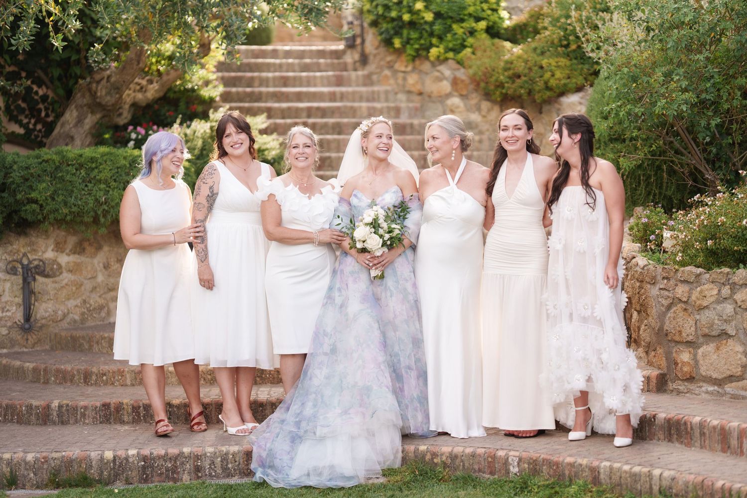 Bride in floral gown poses with six bridesmaids in white dresses on stone steps at outdoor wedding venue.