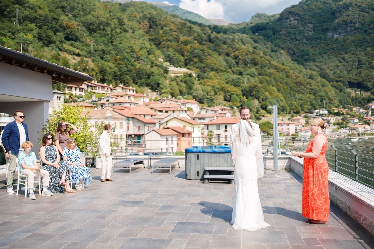 Outdoor wedding ceremony on a lakeside terrace with mountain village backdrop, bride in white gown, officiant in orange dress.