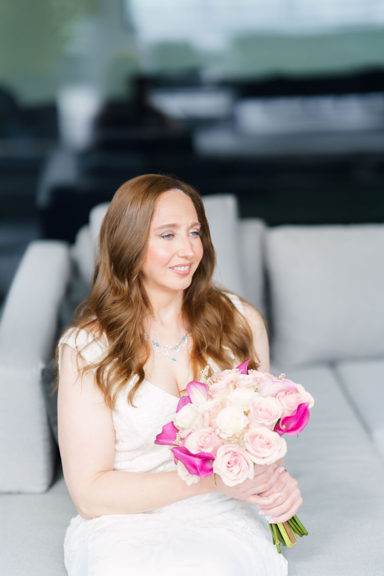 Bride in white dress holding pink and cream rose bouquet, sitting on gray sofa, smiling softly.