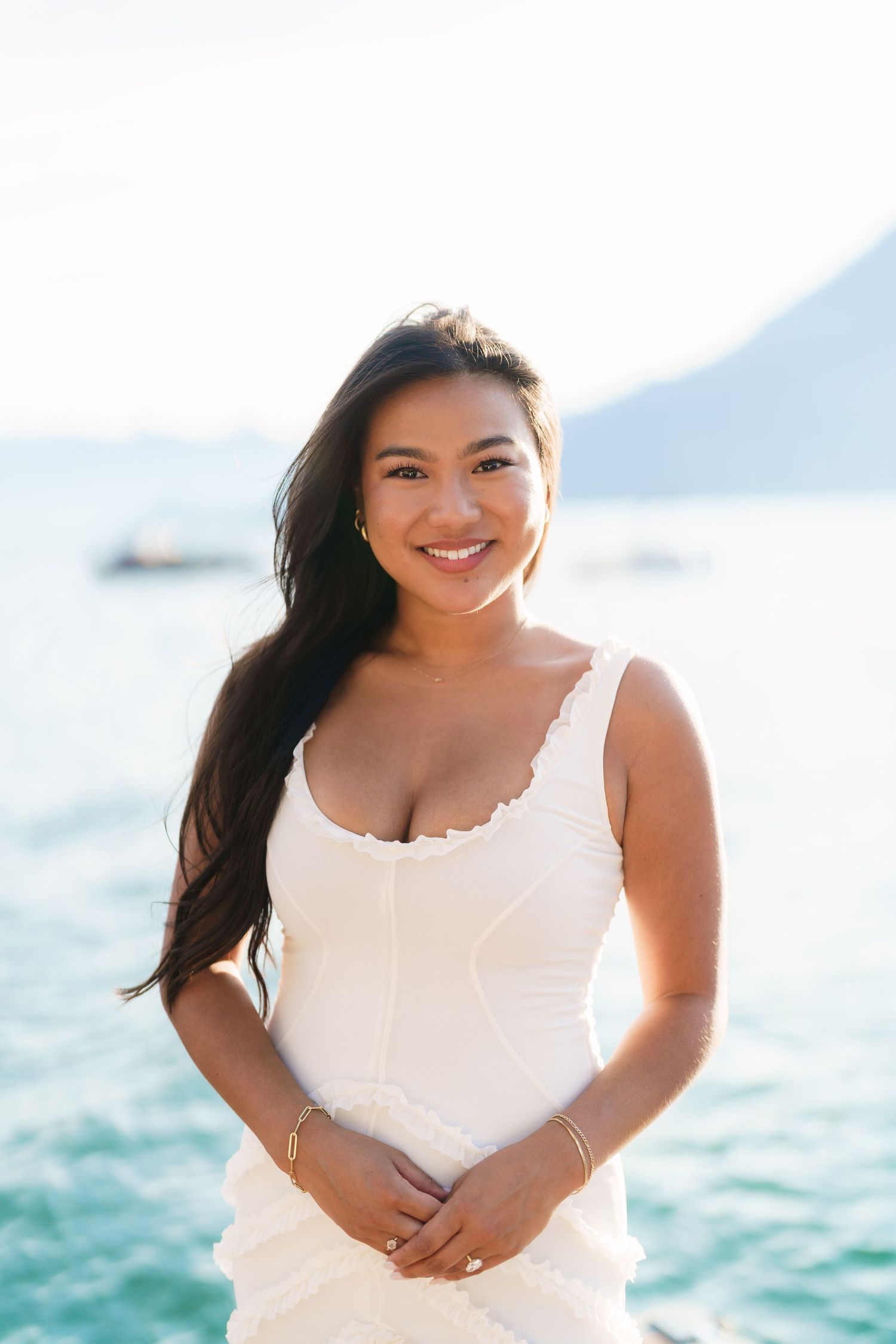 Smiling young woman in white dress posing near ocean with mountains in background on sunny day.