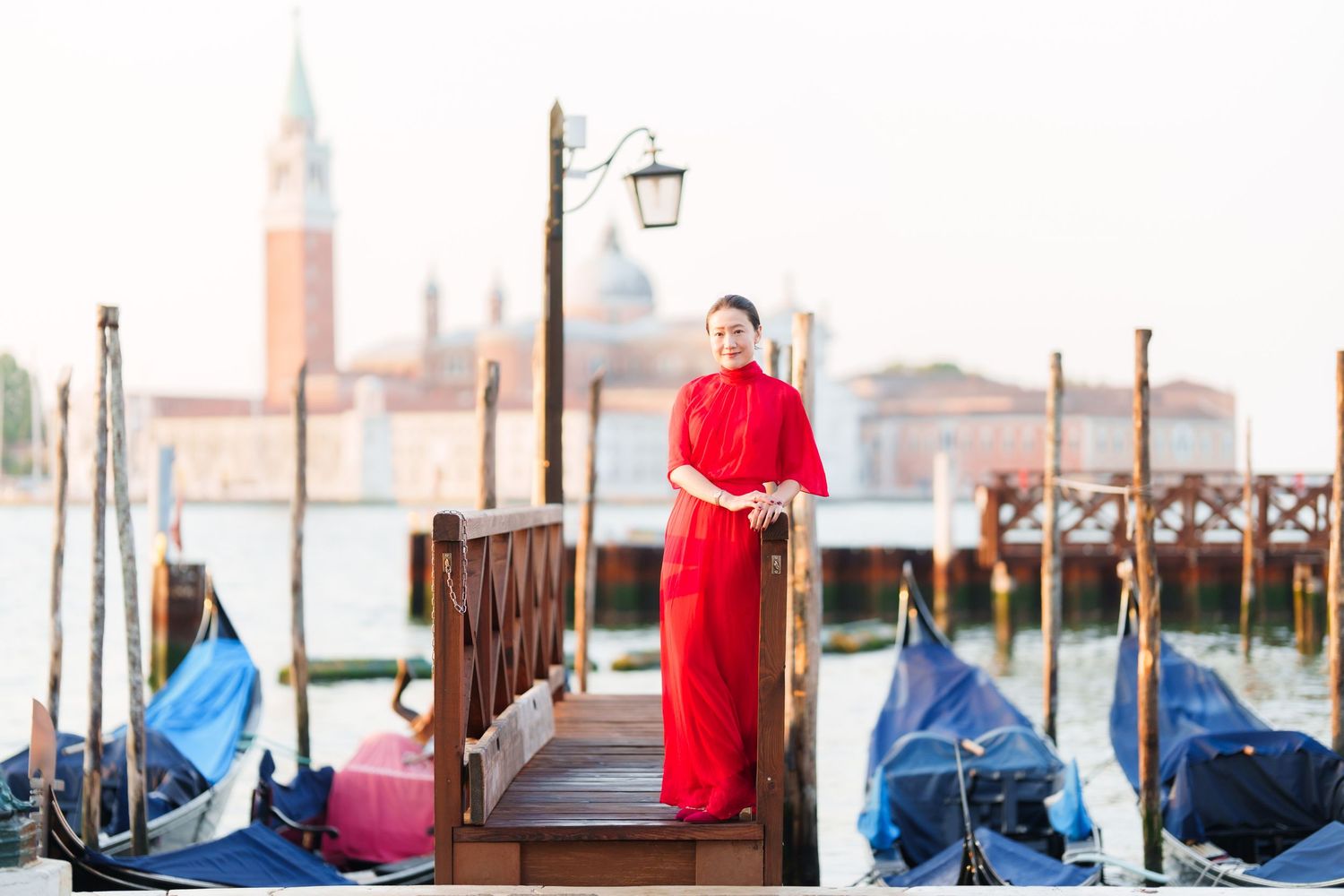 Woman in red dress standing on Venice dock with gondolas and San Giorgio Maggiore church in background.