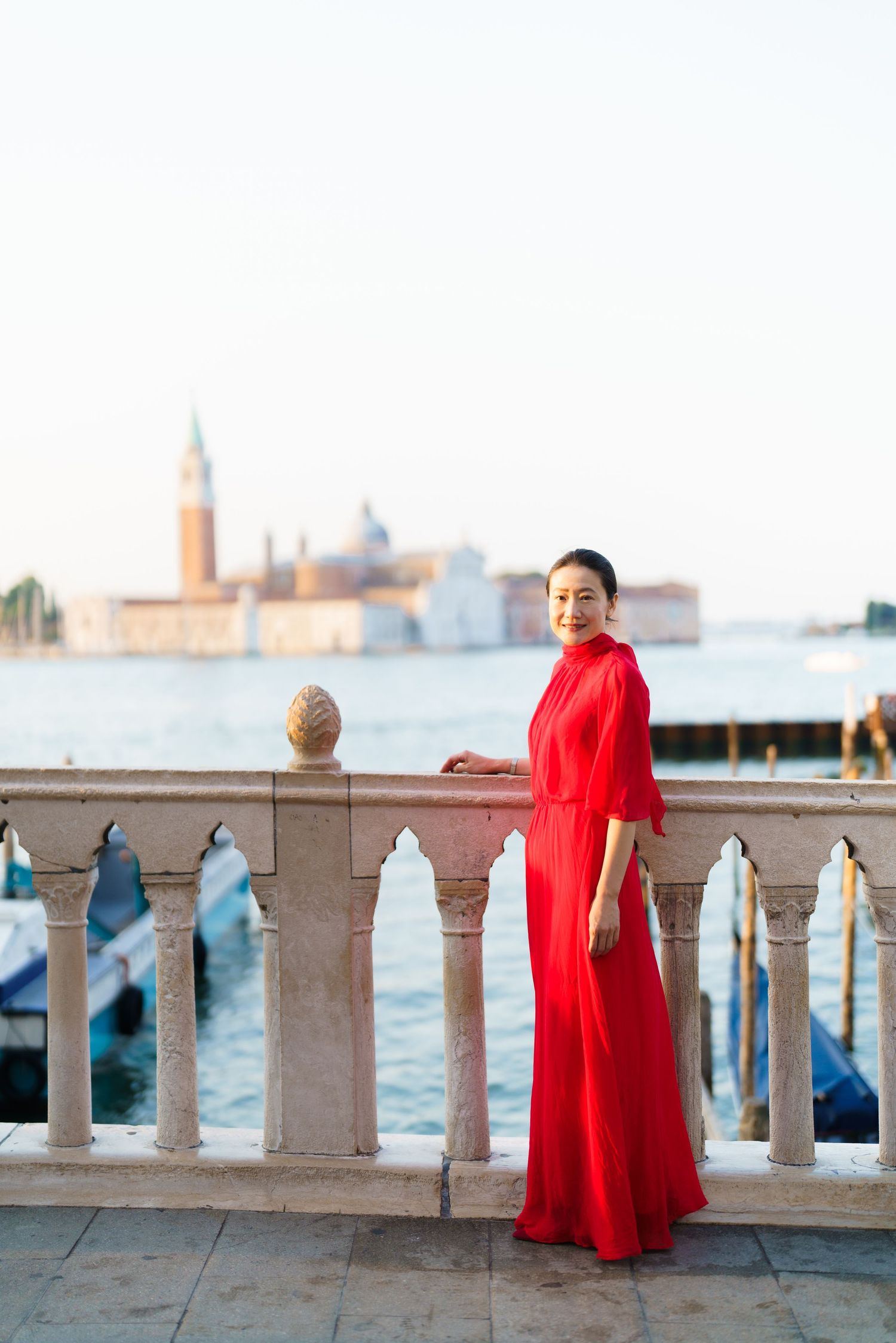 Woman in red dress poses on Venice waterfront balcony with San Giorgio Maggiore island in background.