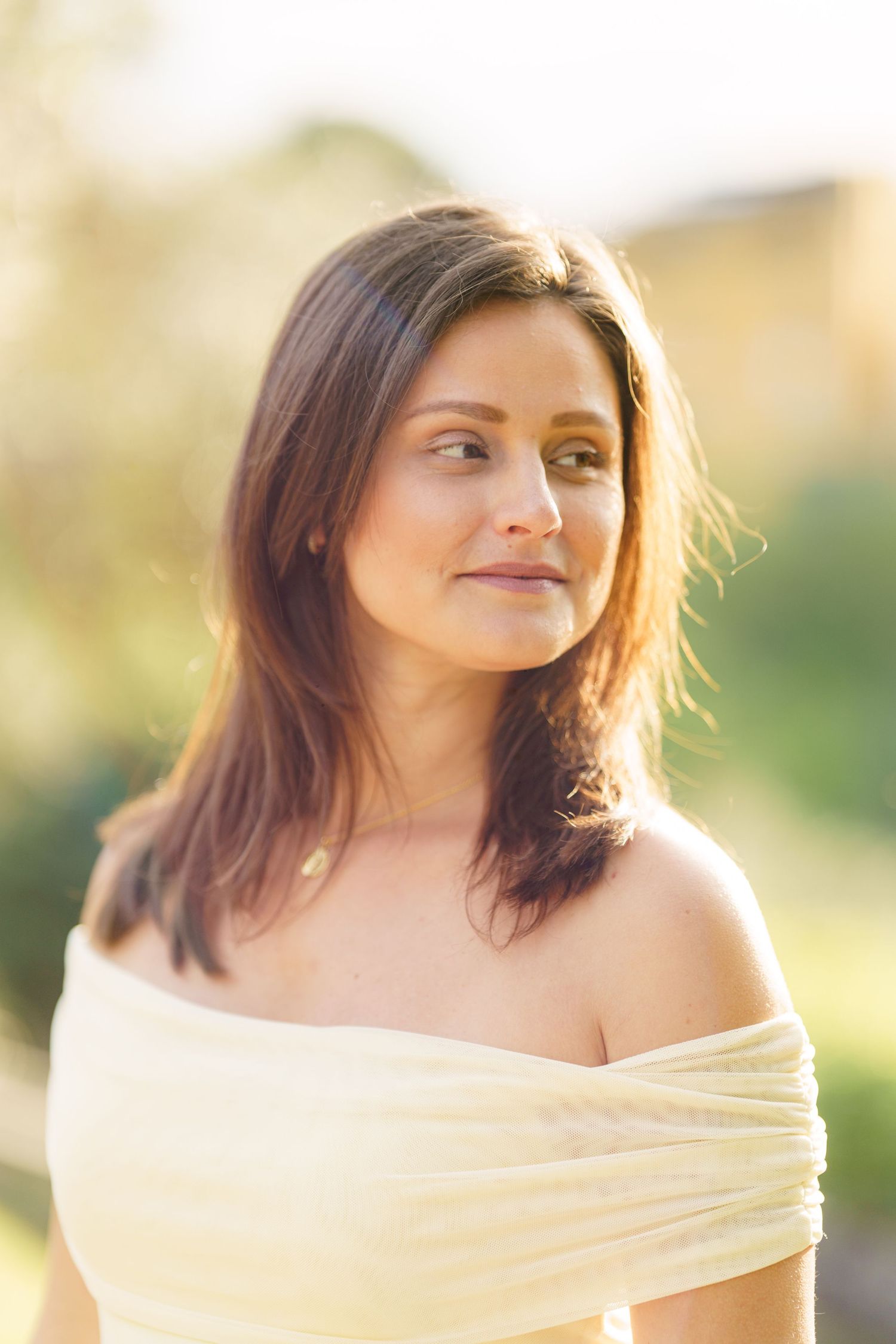 Woman in off-shoulder cream top with brown hair smiling softly outdoors in warm golden sunlight.