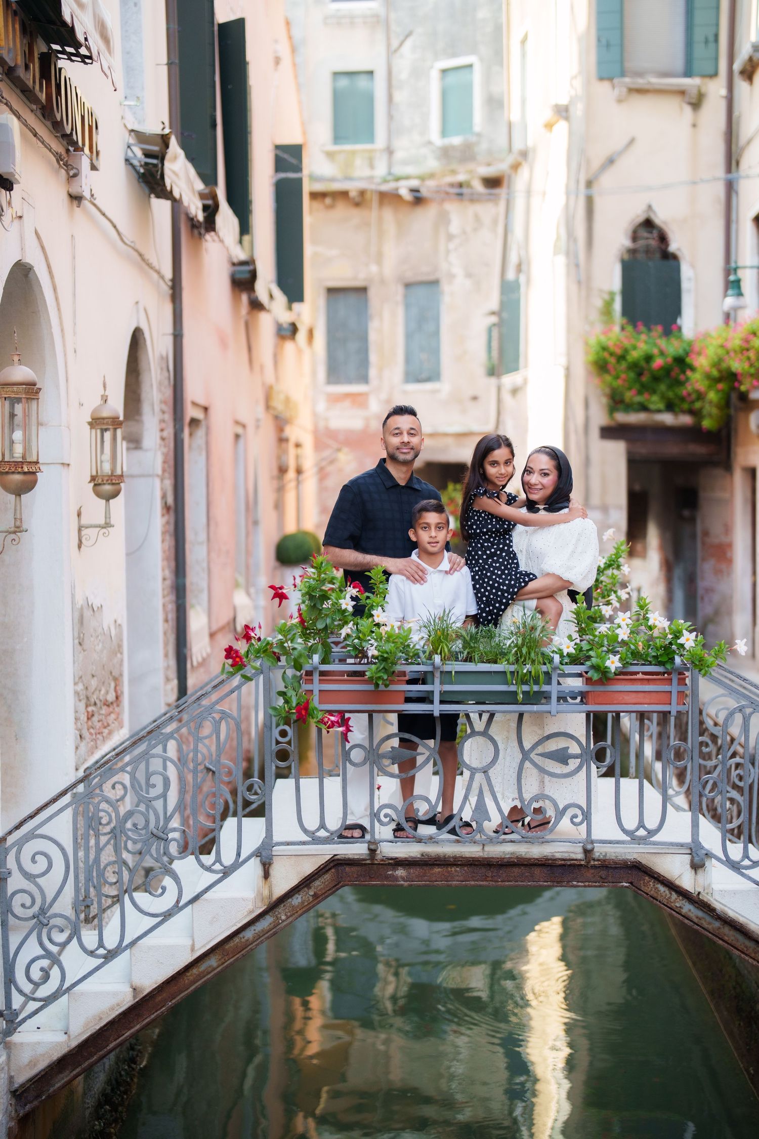 Family of four posing on a charming bridge over a Venetian canal surrounded by colorful flowers in Venice, Italy.