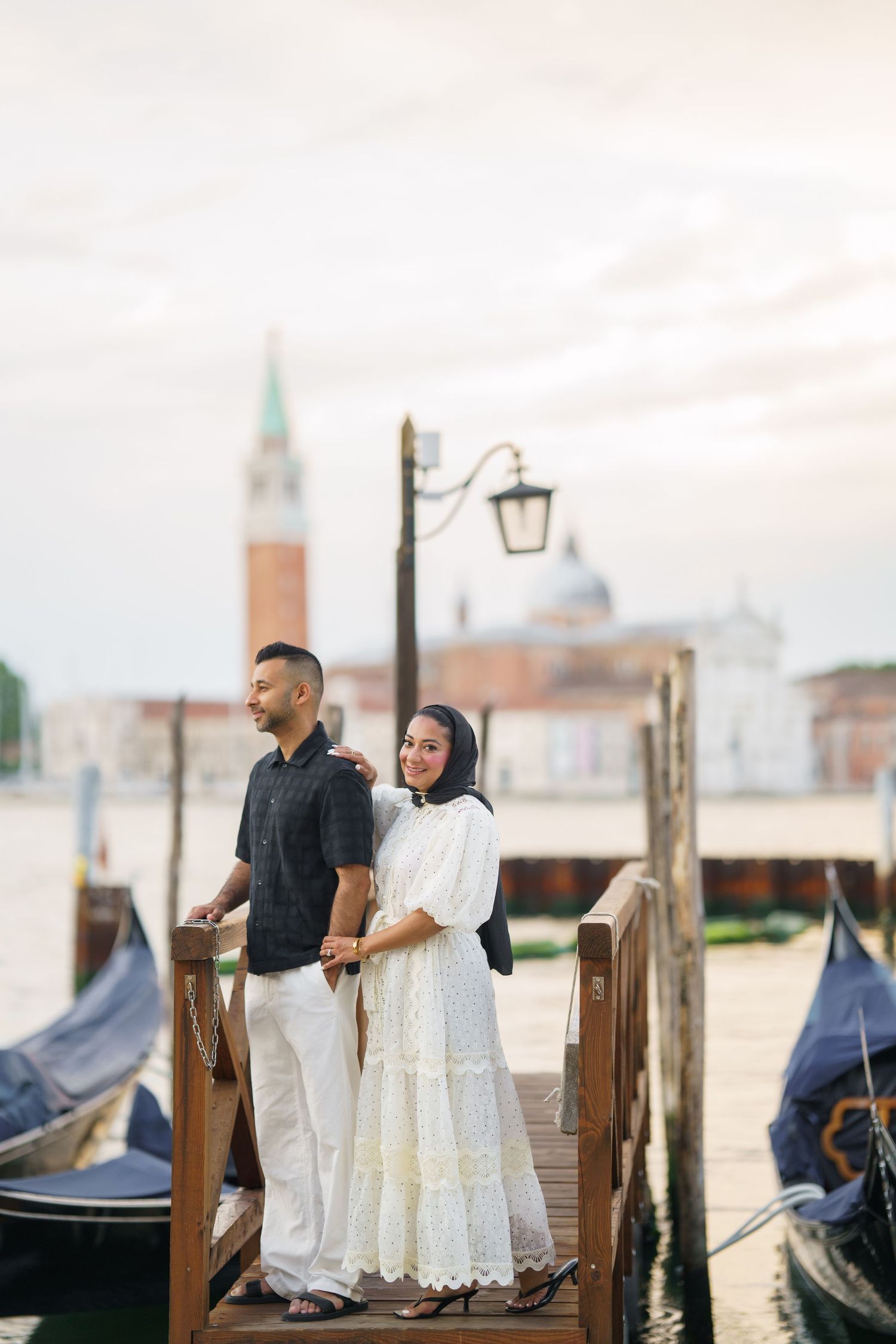 Couple posing on a wooden dock in Venice, Italy, with gondolas and San Giorgio Maggiore church in background.