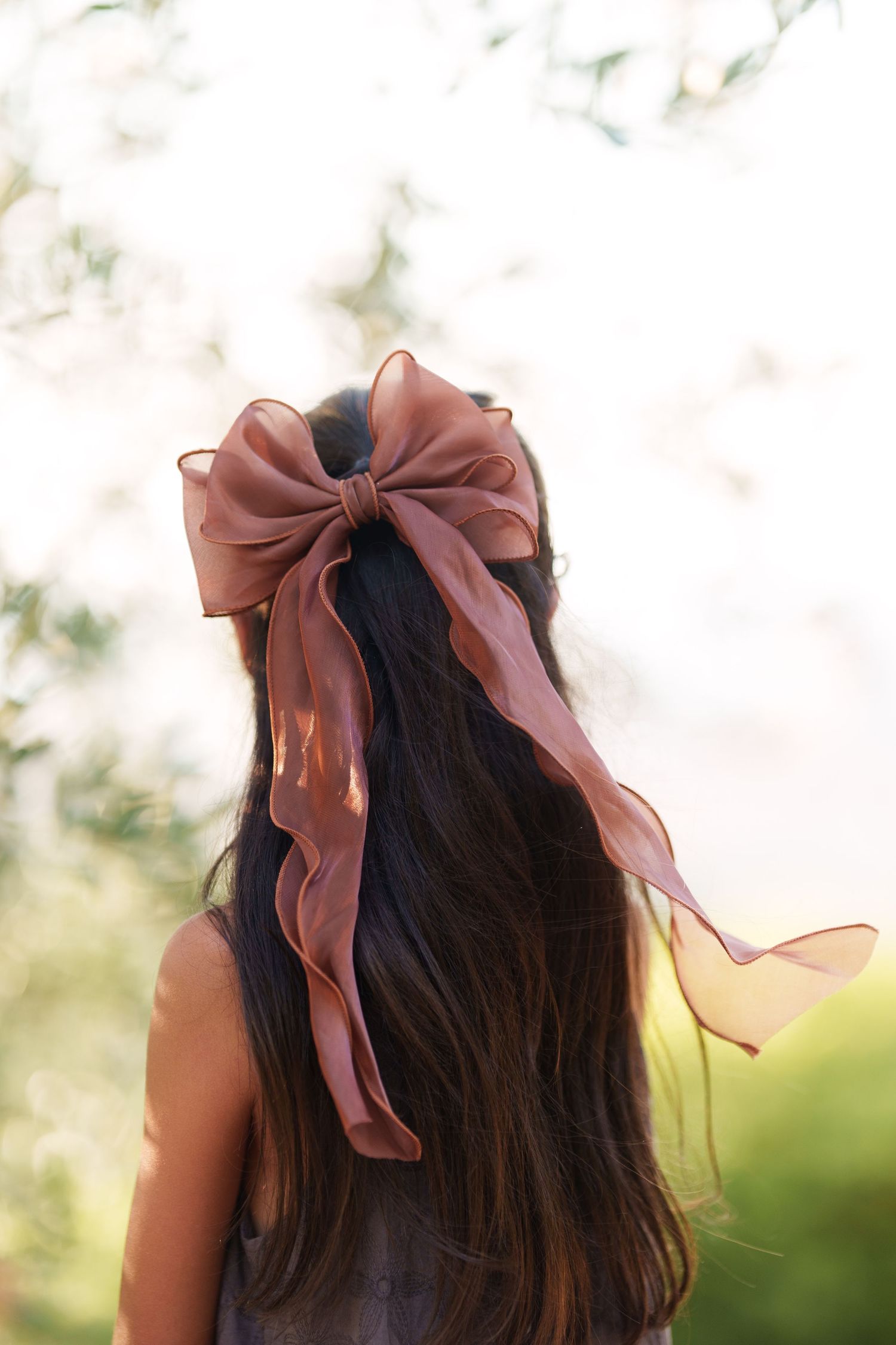 Girl with long dark hair wearing a mauve organza bow hair accessory, photographed outdoors in natural light.
