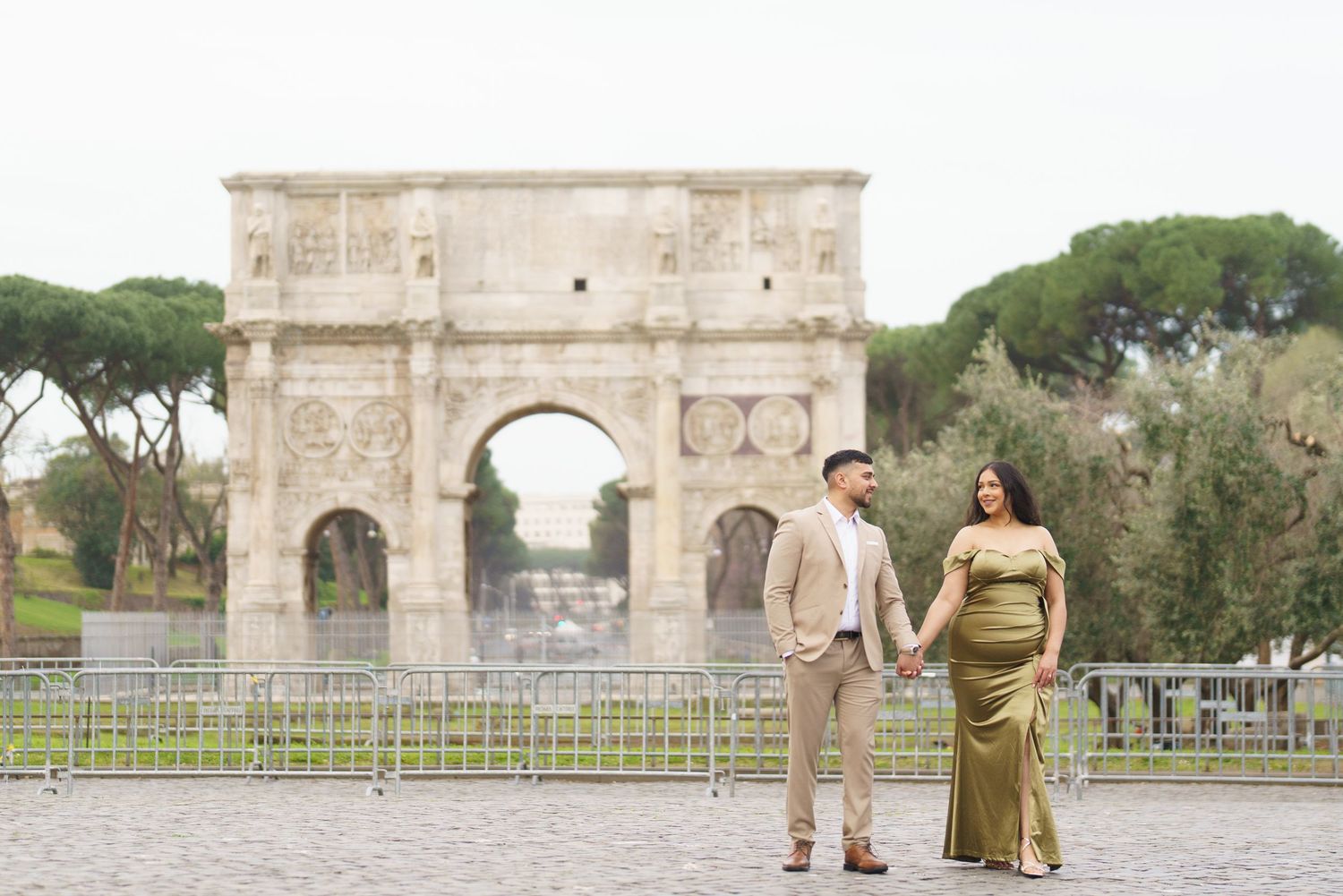 Couple holding hands in front of the Arch of Constantine in Rome, Italy, wearing elegant attire.