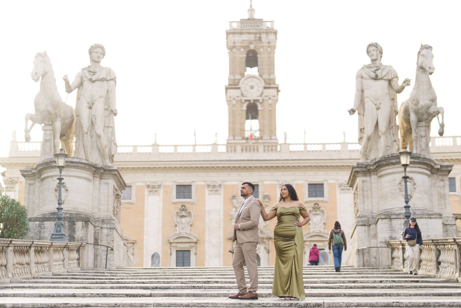 Couple posing on Capitoline Hill steps in Rome, Italy, with classical statues and historic Palazzo Senatorio backdrop.