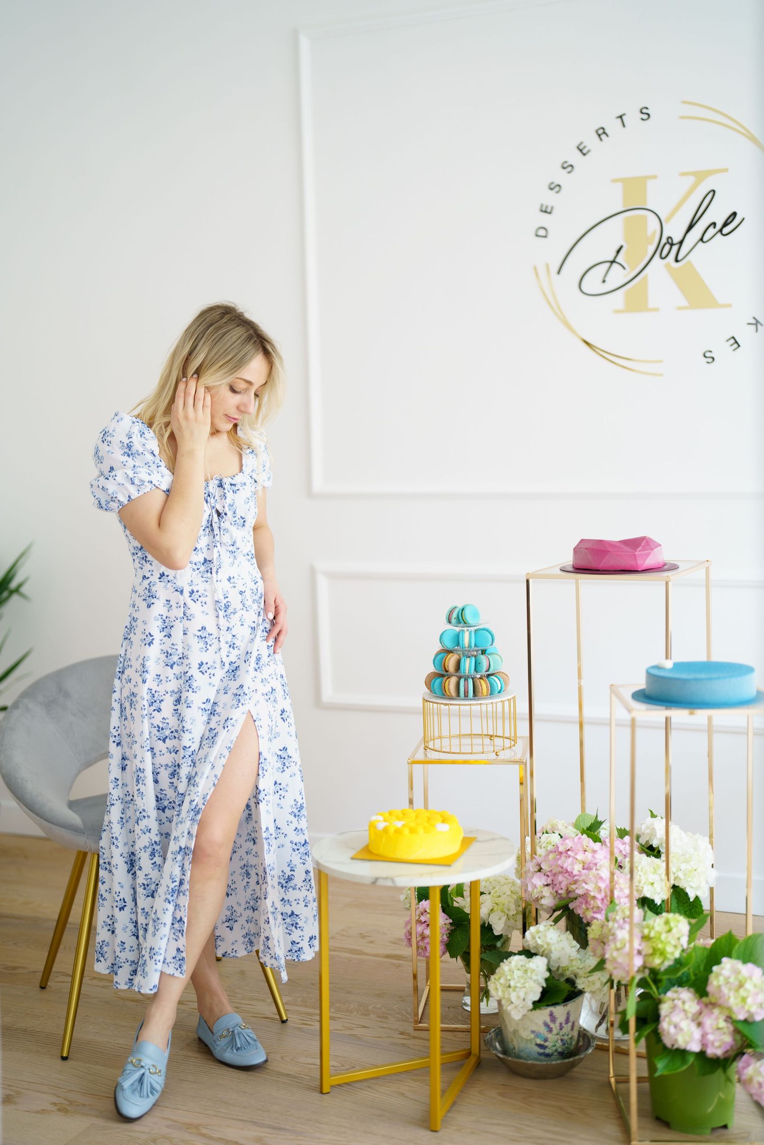Woman in floral dress poses near elegant dessert display at Dolce K Desserts boutique with macarons and cakes.