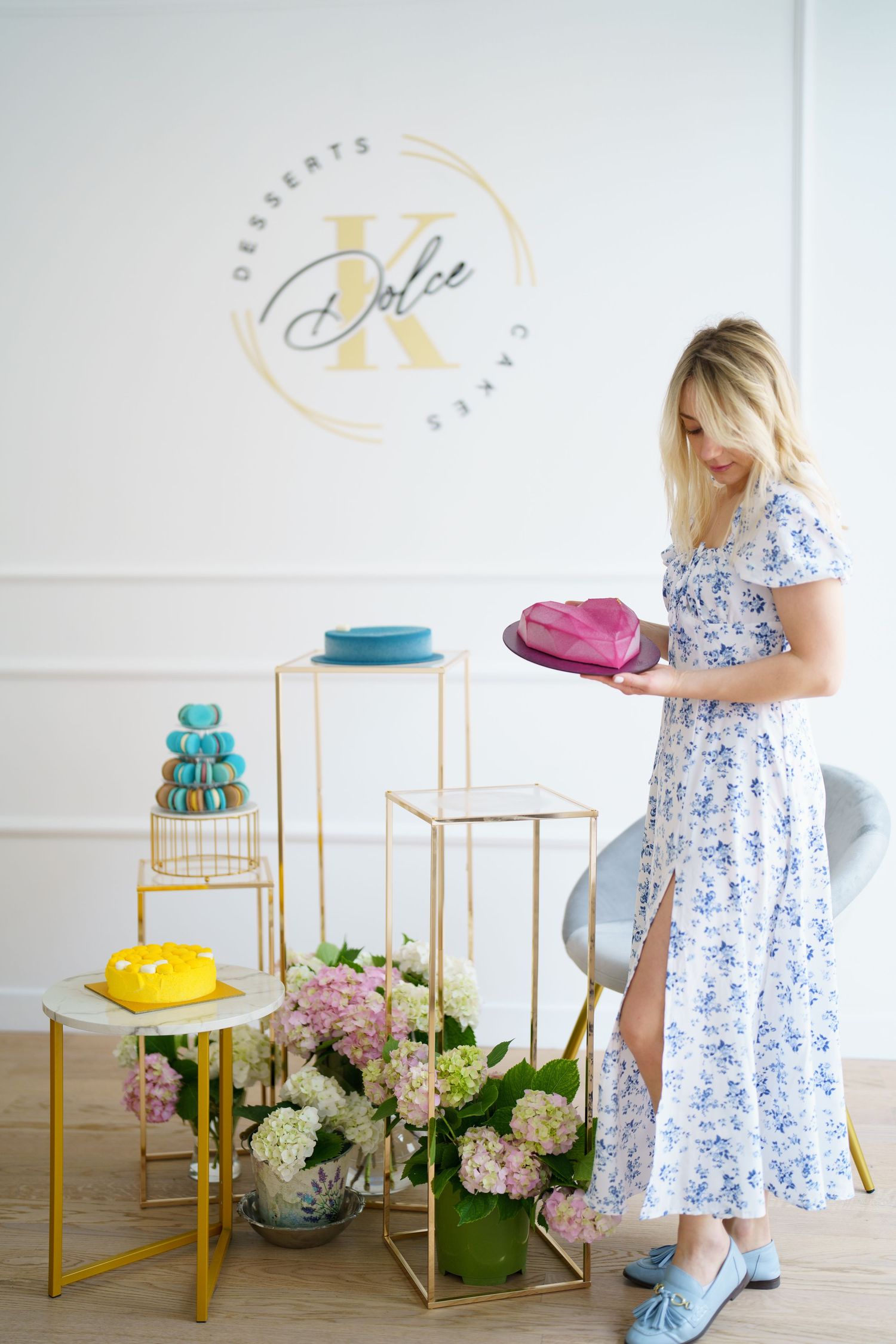 Woman in floral dress holding pink cake at Dolce Desserts bakery with macarons and hydrangeas display.
