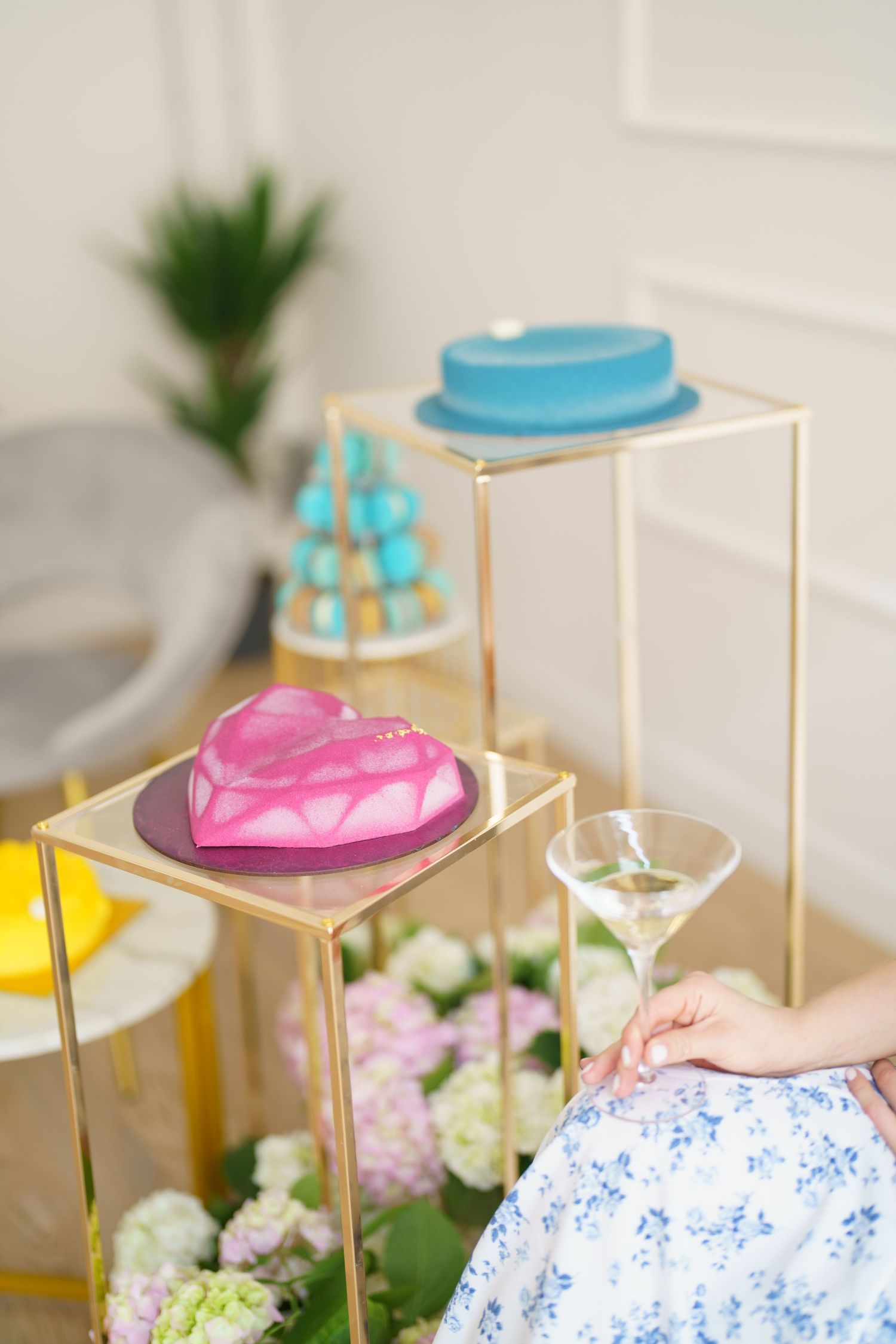 Elegant dessert display with pink heart cake and blue round cake on gold stands, with martini glass and flowers.