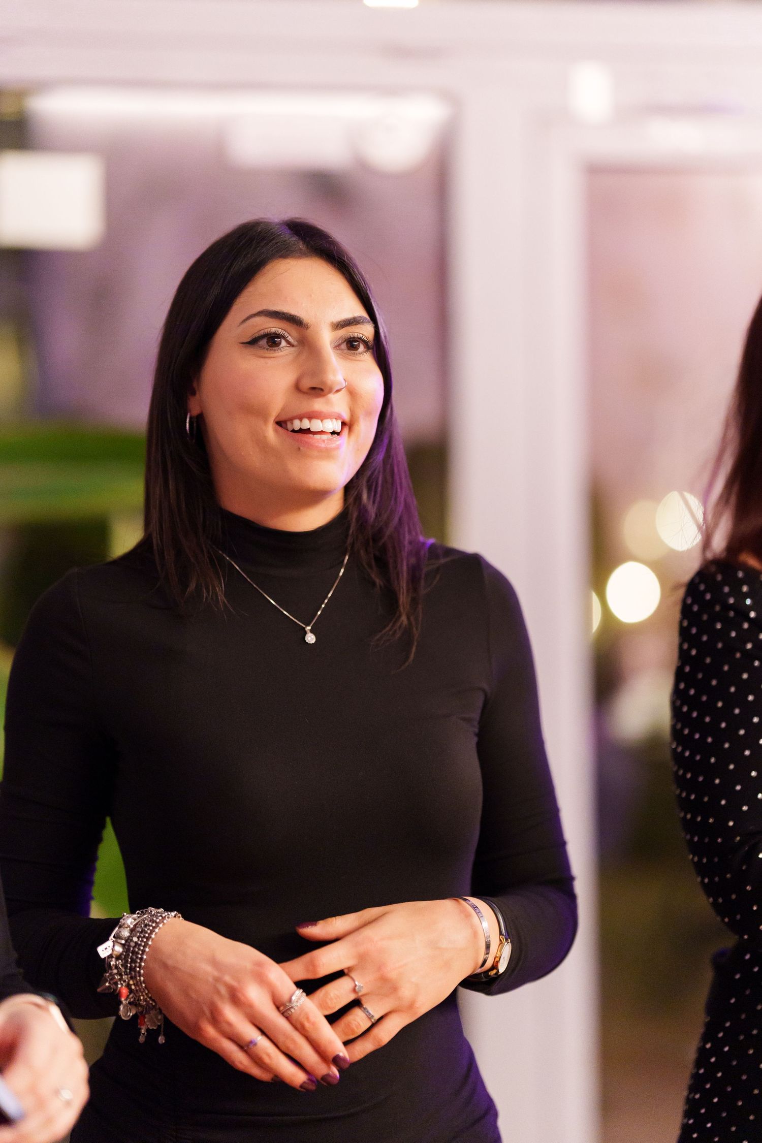 Smiling woman in black turtleneck with silver jewelry at an indoor event.