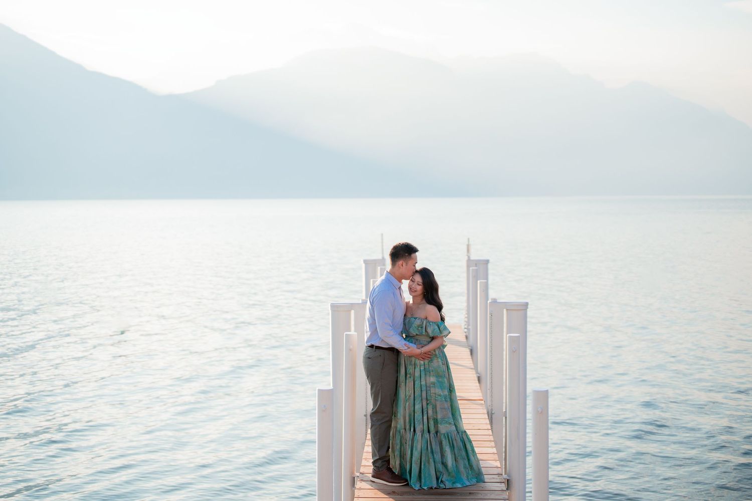 Couple embracing on a wooden dock overlooking a serene mountain lake at dusk, woman in teal gown.