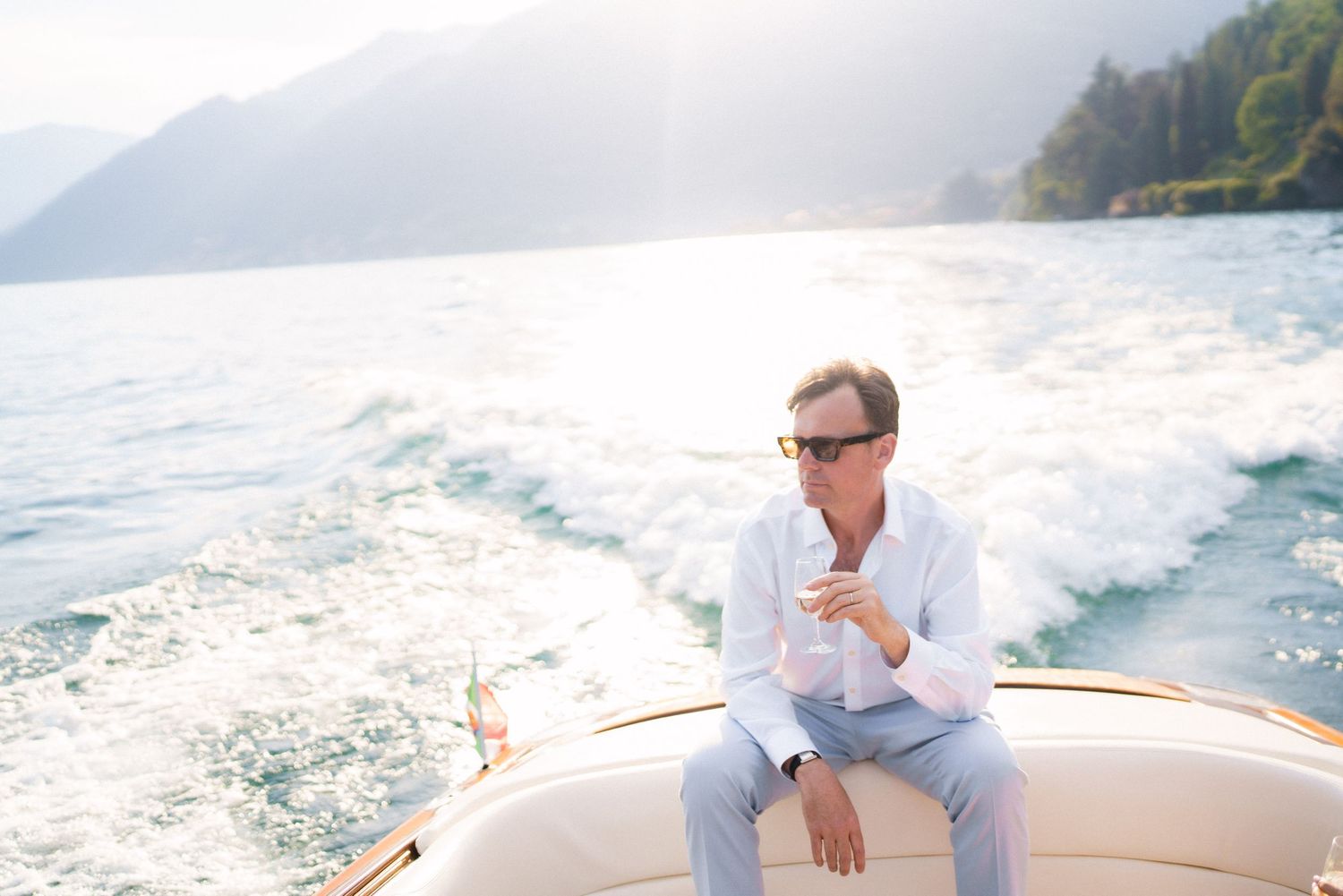 Man in white shirt holding a drink on a luxury boat with mountain lake scenery in the background.