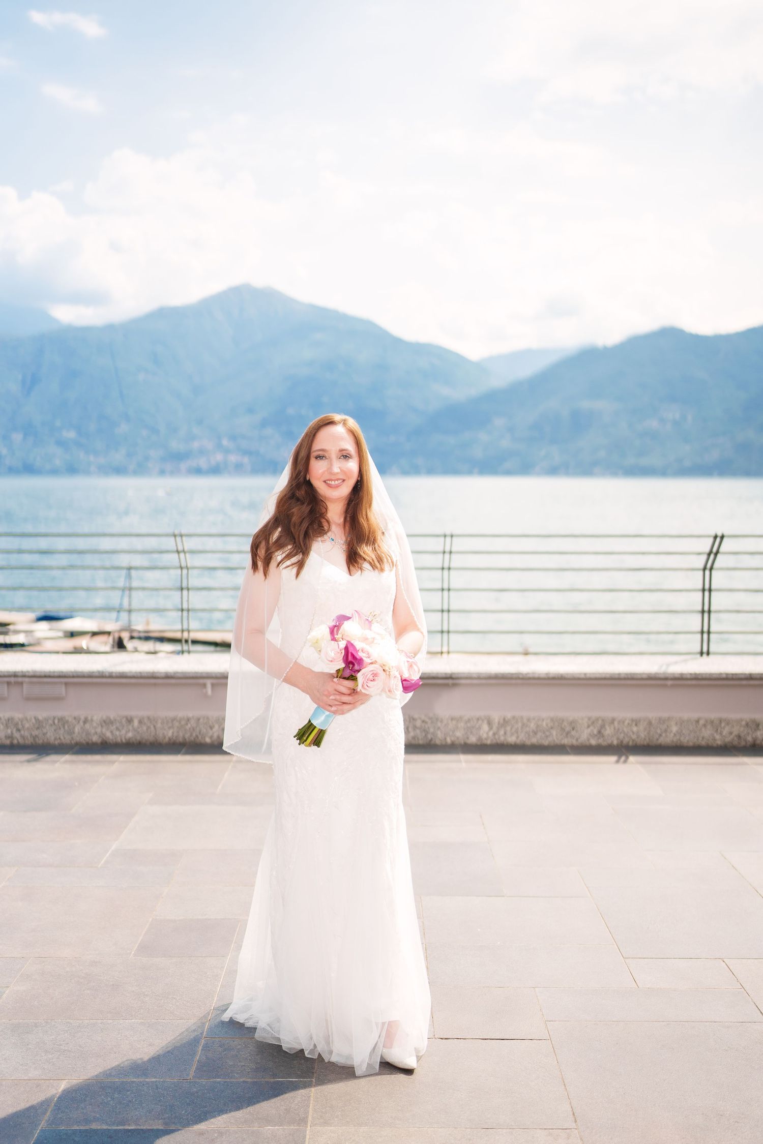 Bride in white gown and veil holds pink bouquet on terrace with mountain lake backdrop como lake