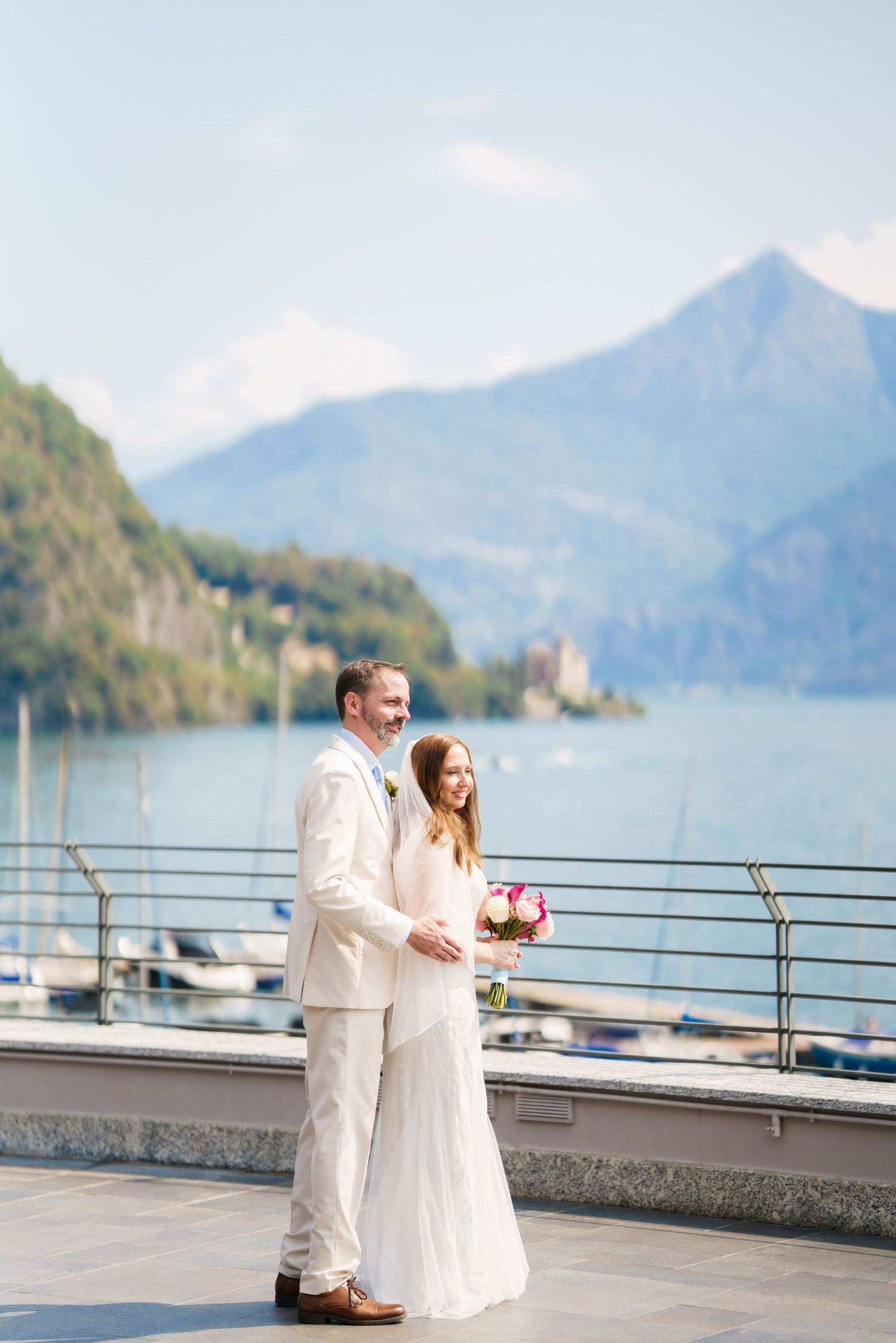Bride and groom standing on a lakeside terrace with mountain views, bride holding pink bouquet, both in white attire.