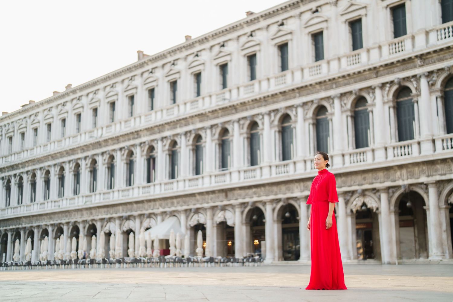 Woman in elegant red gown standing in St. Mark's Square, Venice, Italy, with historic arcade building backdrop.