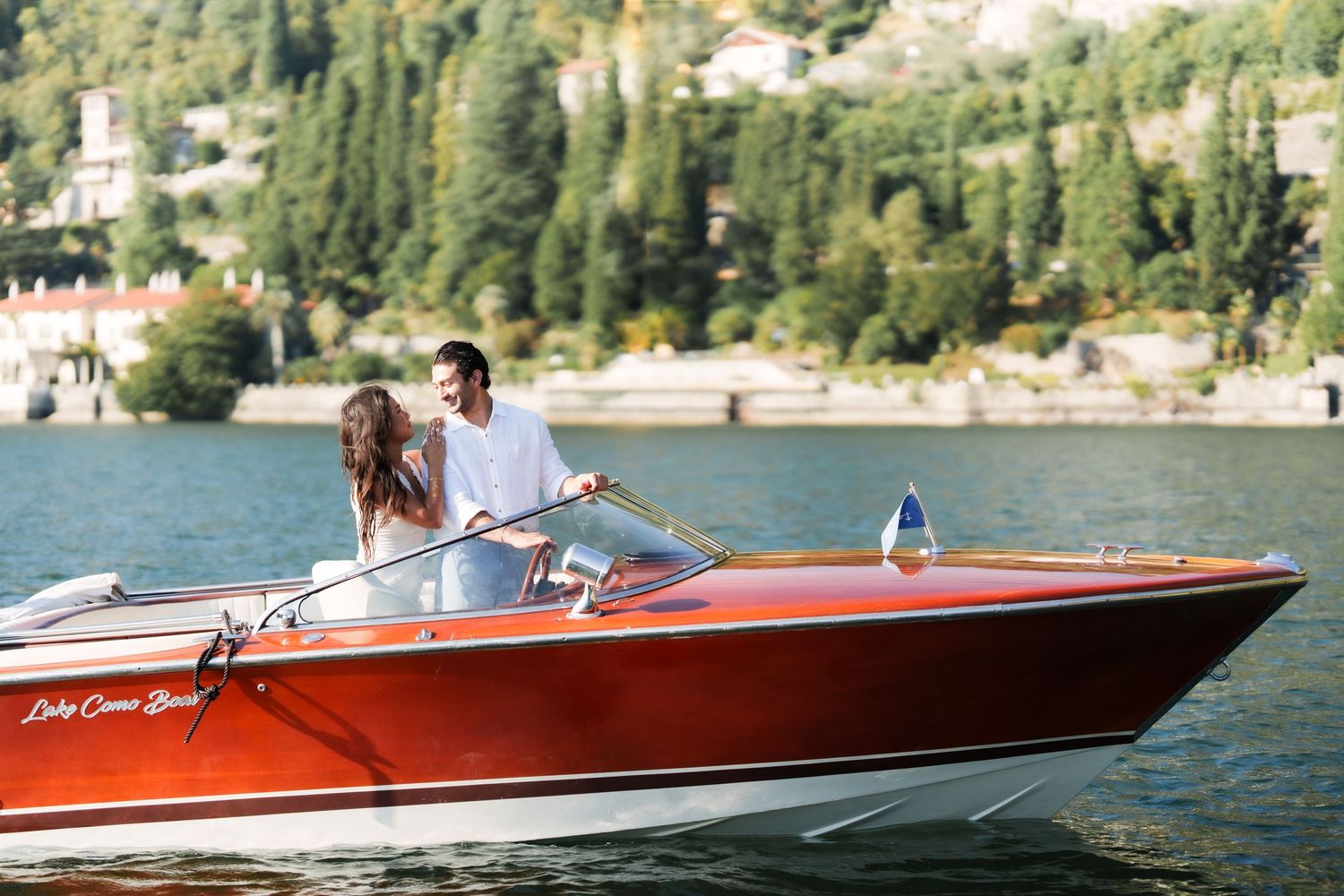 Couple enjoying a romantic boat ride on Lake Como in a classic red wooden speedboat surrounded by lush green hills.