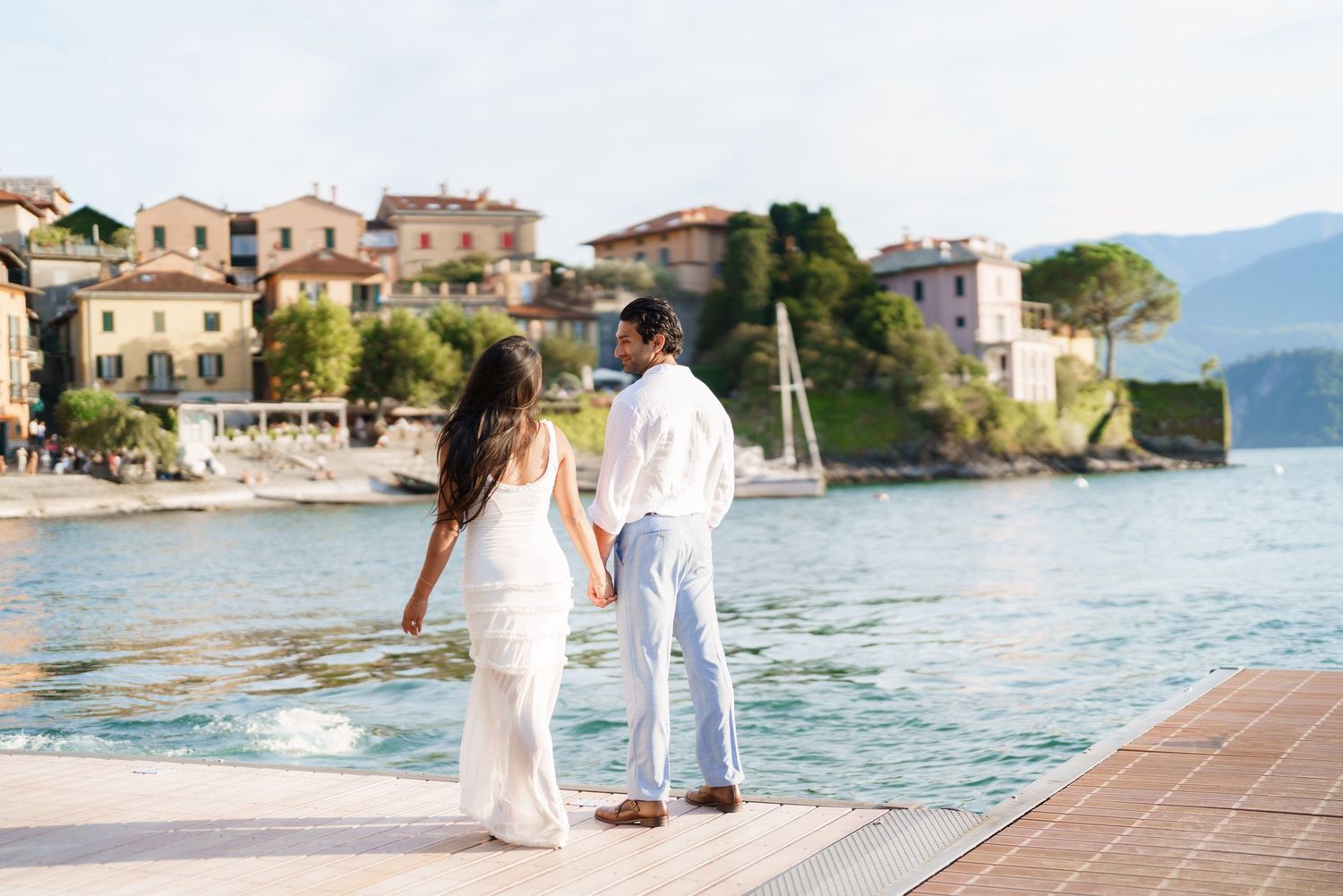 Couple in wedding attire holding hands on a dock overlooking a scenic Italian lake village with colorful buildings.