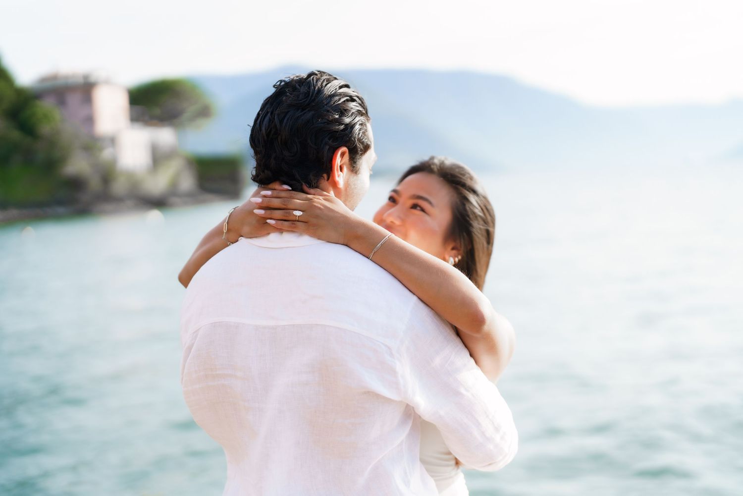 Couple embracing romantically by a serene lake with mountains and buildings in the background.