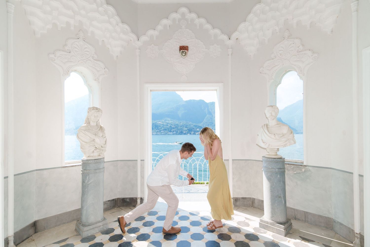 Man proposing to woman in elegant white marble hall with arched windows overlooking scenic lake and mountains.