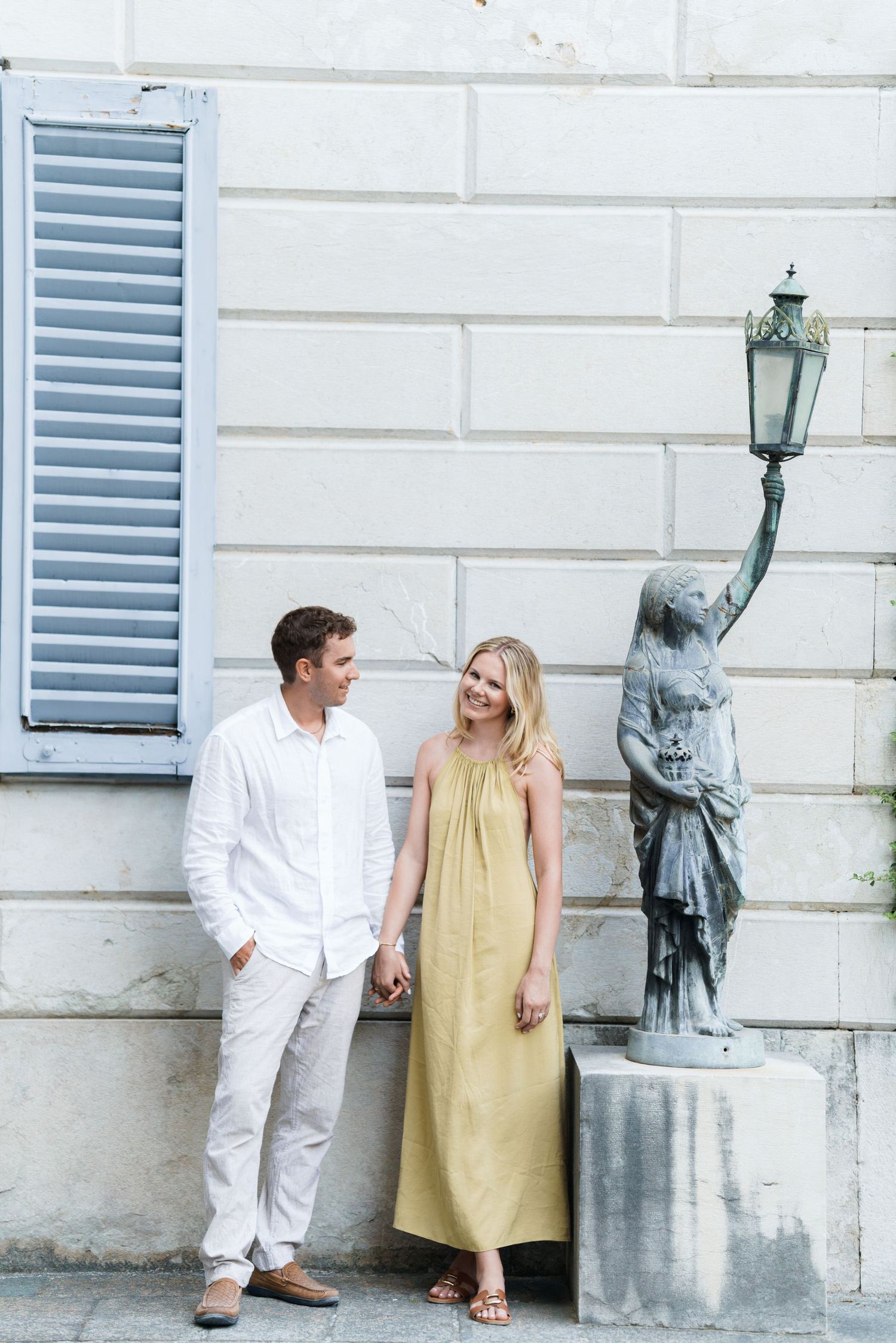 Couple in white and yellow outfits holding hands beside a statue lamppost against a white stone wall.