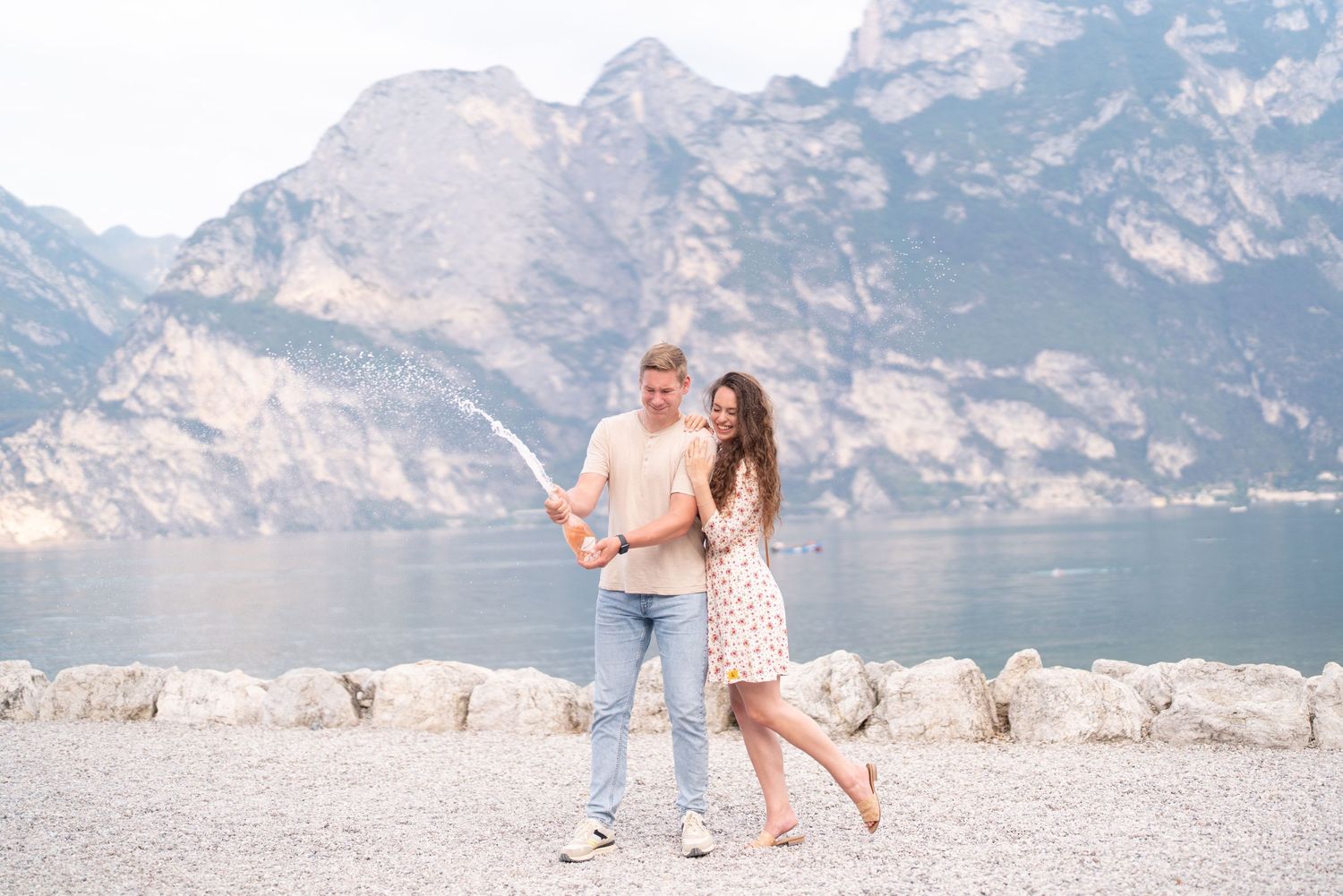 Couple smiling and holding a map by a scenic mountain lake with rocky shoreline in the background.