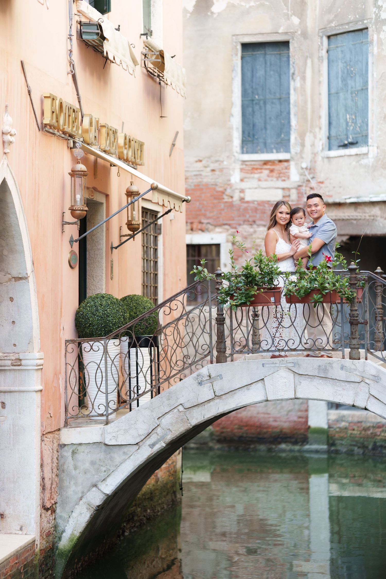 Family posing on a charming stone bridge over a Venice canal, surrounded by colorful buildings and lush greenery.