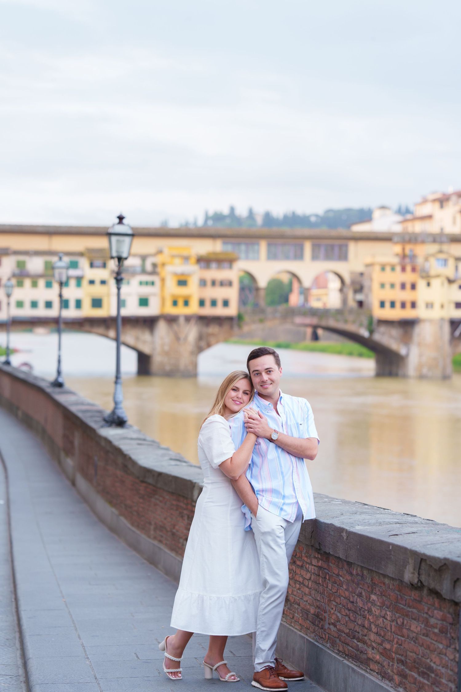 Romantic couple in white outfits posing along the historic Ponte Vecchio bridge in Florence, Italy at sunset.