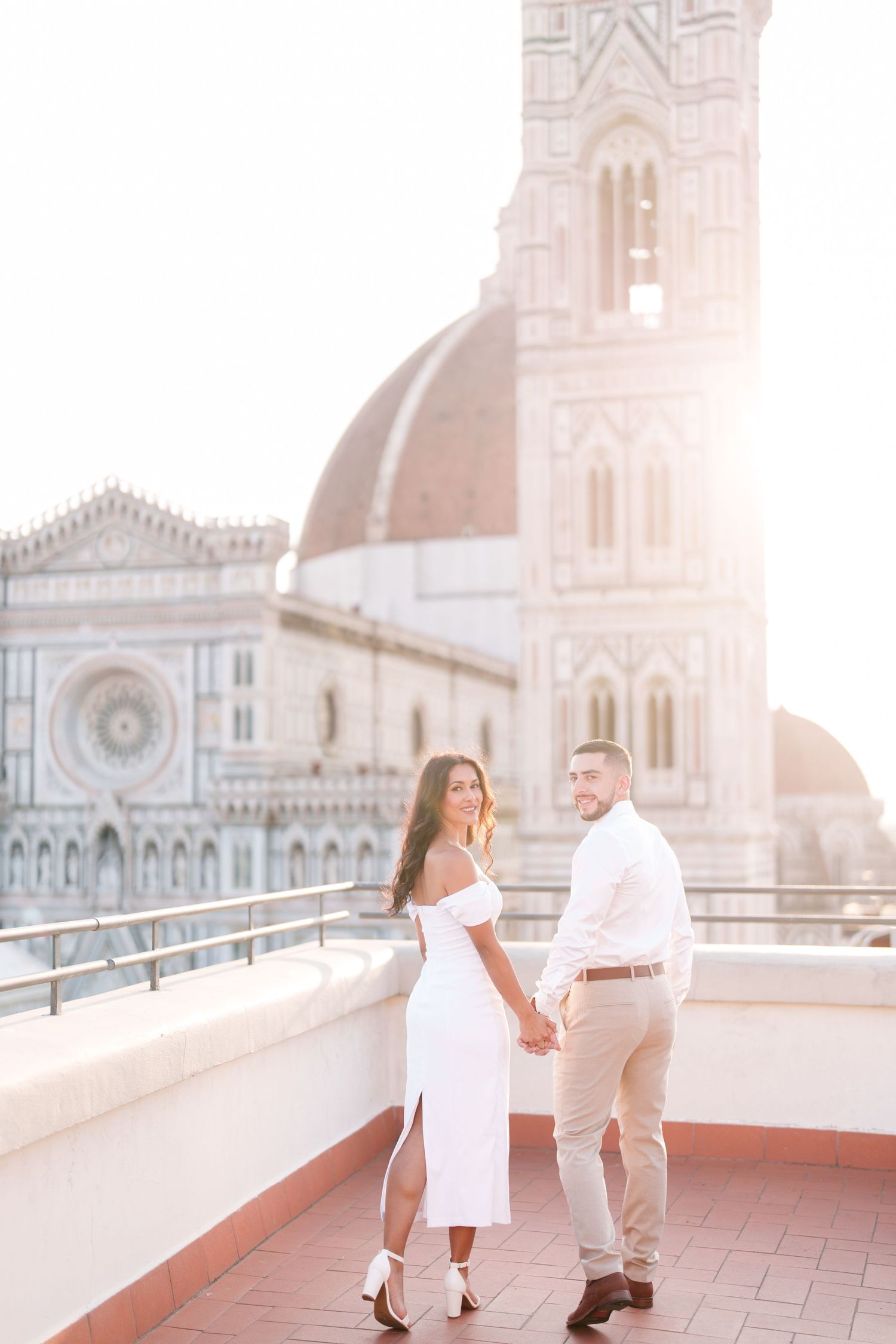 A couple in white outfits poses on a rooftop terrace with Florence Cathedral's iconic dome and bell tower in the background.