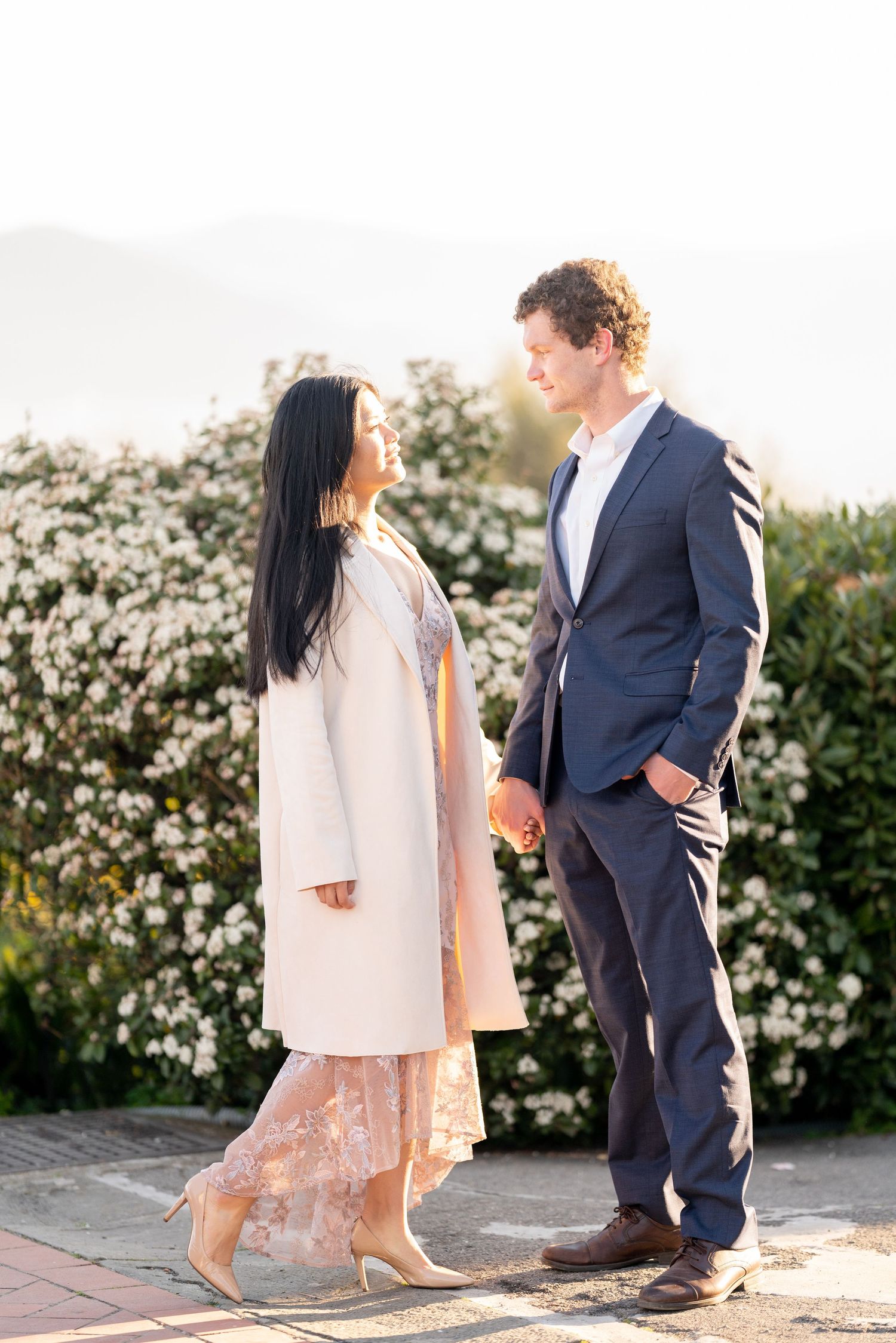 A couple in elegant attire stands together against a backdrop of blooming white flowers during golden hour.
