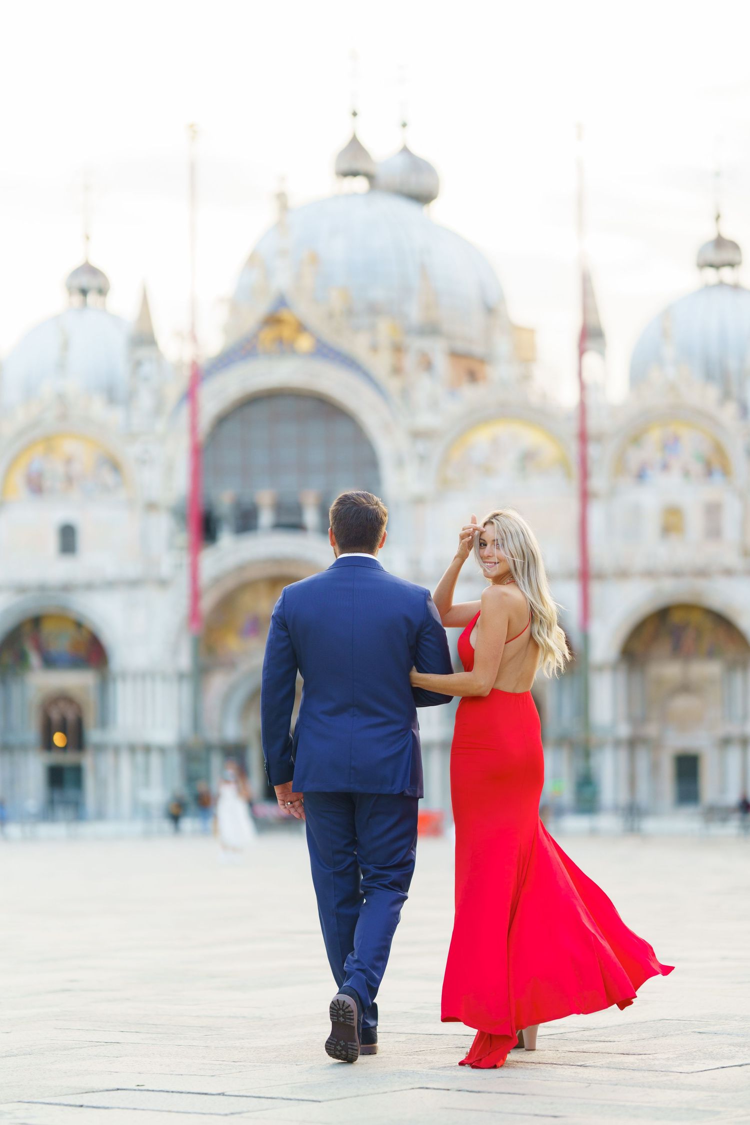 A couple in formal attire walks through St. Mark's Square in Venice with the basilica's ornate facade in the background.