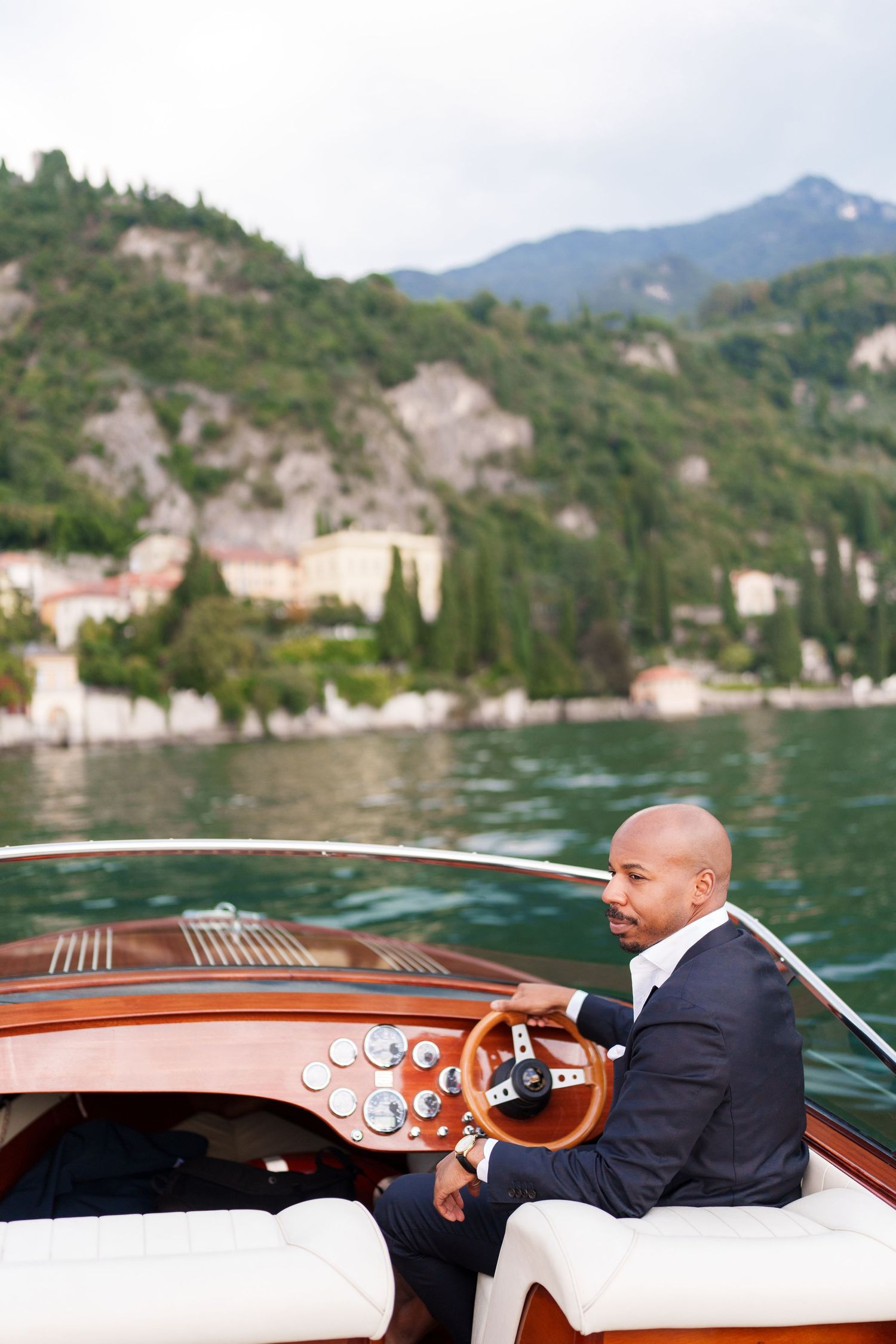 A person in a suit steers a luxury wooden boat across a scenic mountain lake with cliffs in the background.