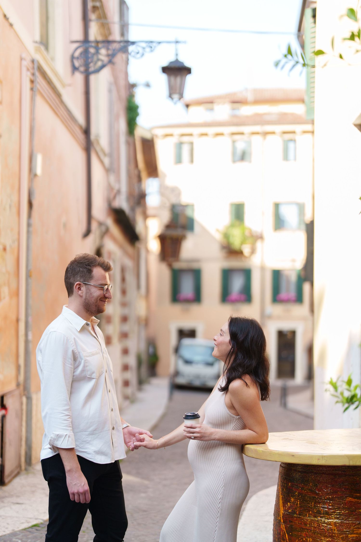 A couple shares a romantic moment while holding drinks on a charming European street with warm colored buildings.